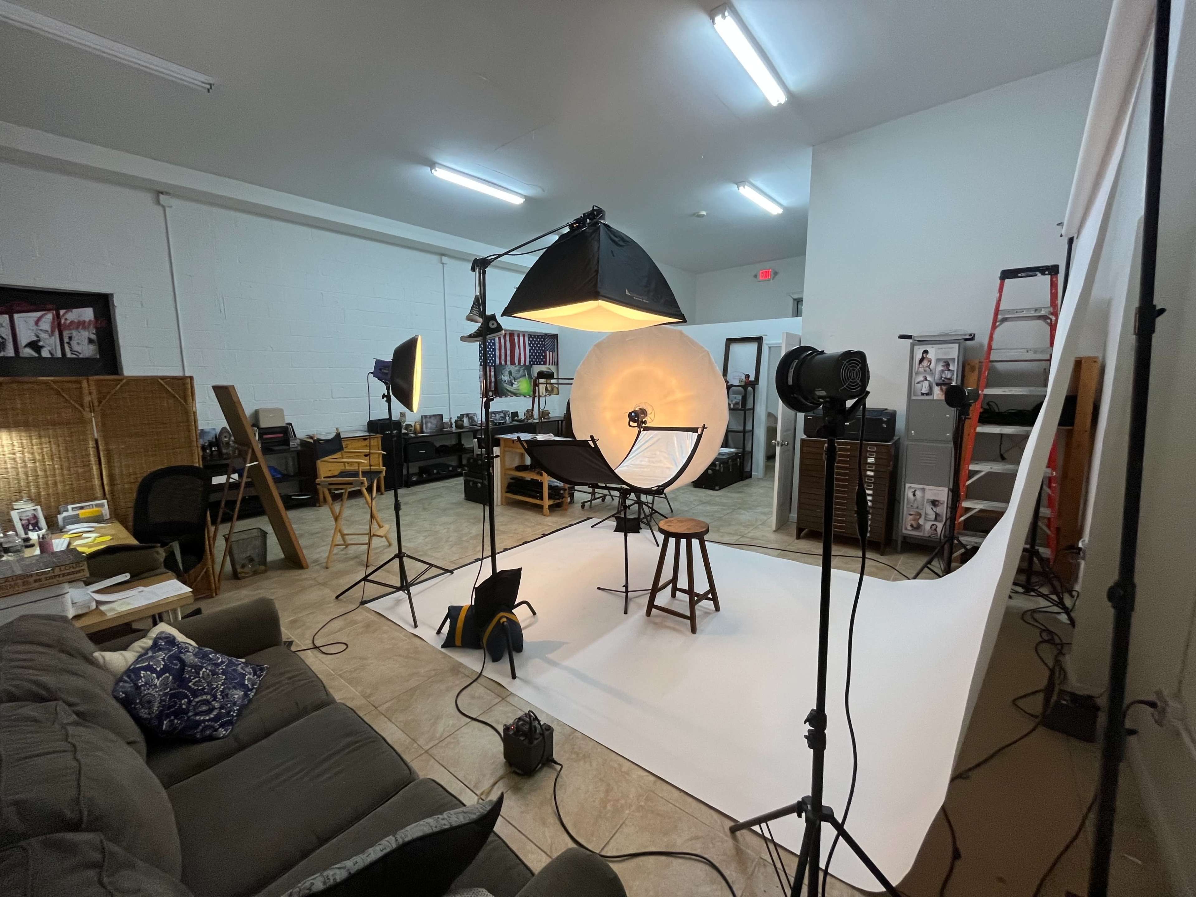 A photography studio set up with various lights, camera equipment, and a stool on a white backdrop, along with a seating area in the corner.
