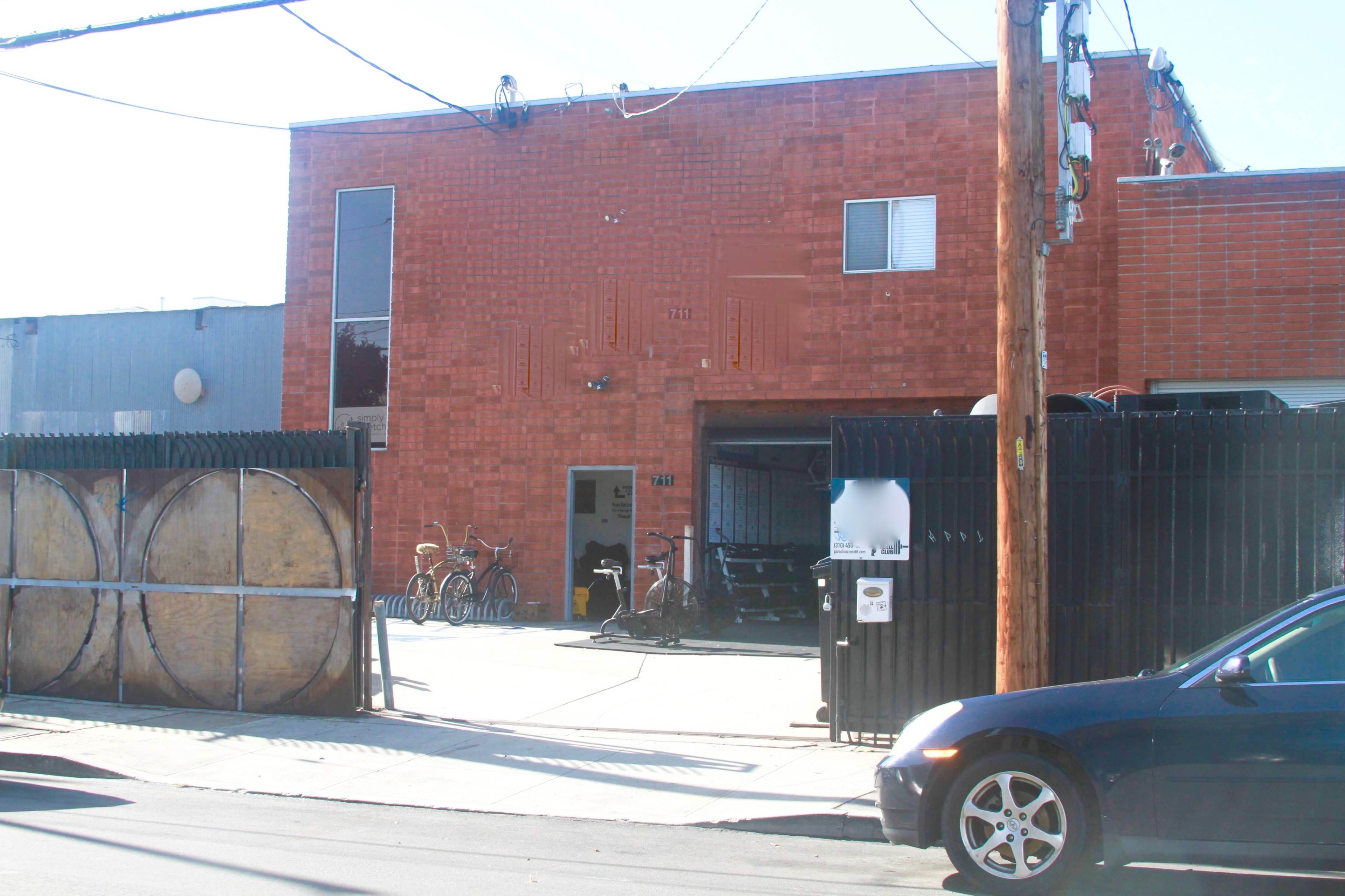 The image shows a red brick building with a large garage door, surrounded by a fenced area containing bicycles and wooden barrels, along with a parked blue car on the street.