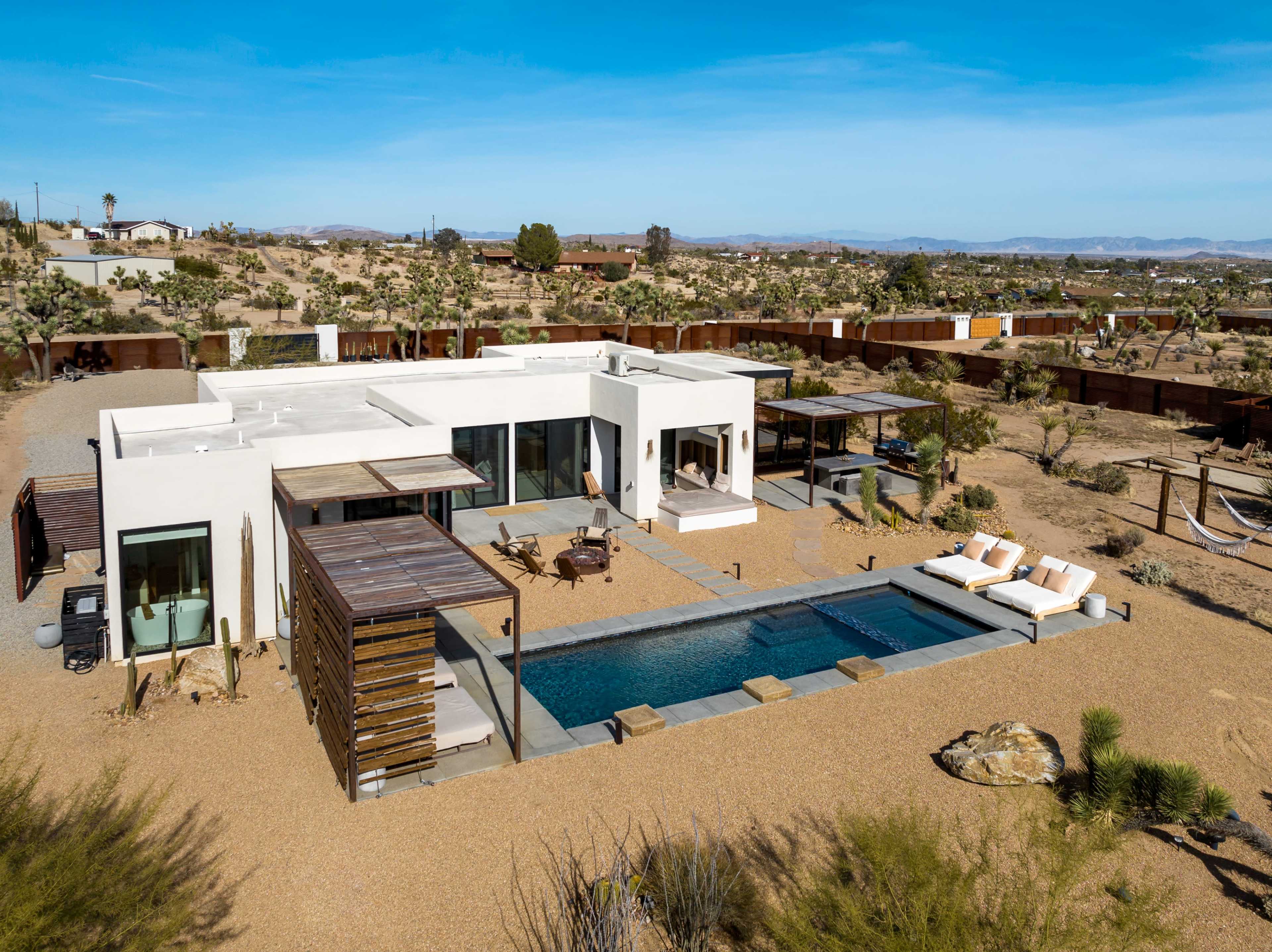 A modern desert villa with a swimming pool surrounded by landscaping, located in a vast arid landscape with distant hills.
