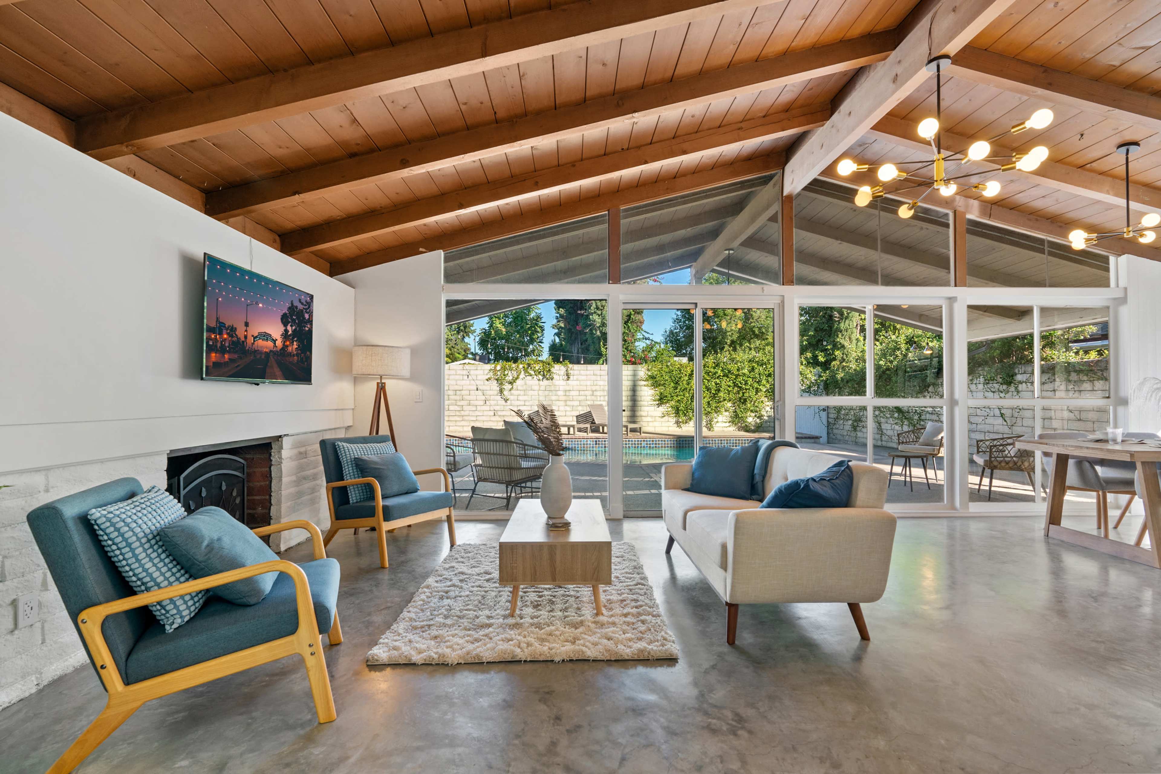 A modern living room with wooden beams, a concrete floor, and large windows overlooking a pool area, featuring two armchairs and a light-colored sofa around a coffee table.