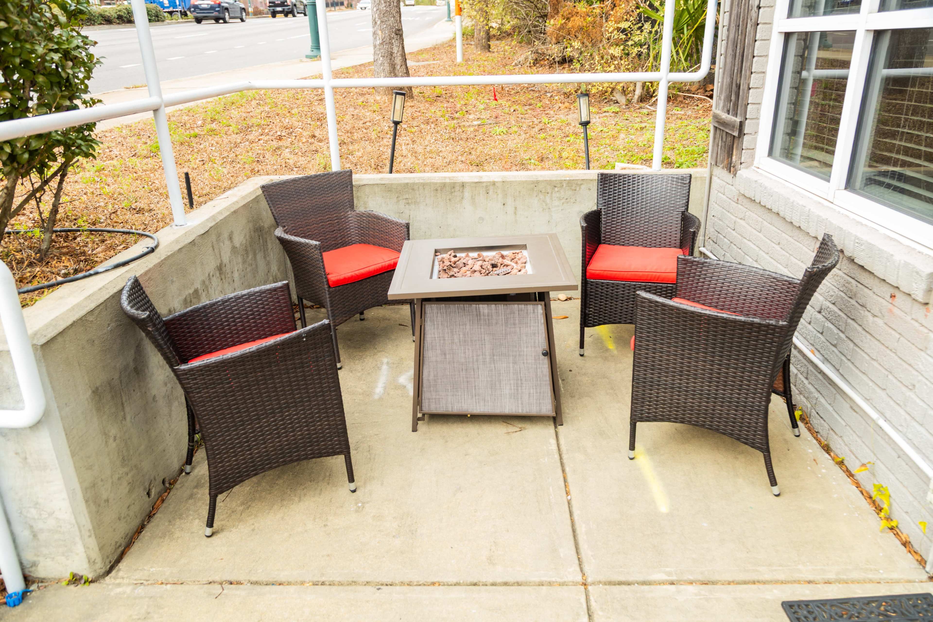 The image shows a small outdoor seating area with four wicker chairs and a fire pit table in the center, surrounded by concrete walls.