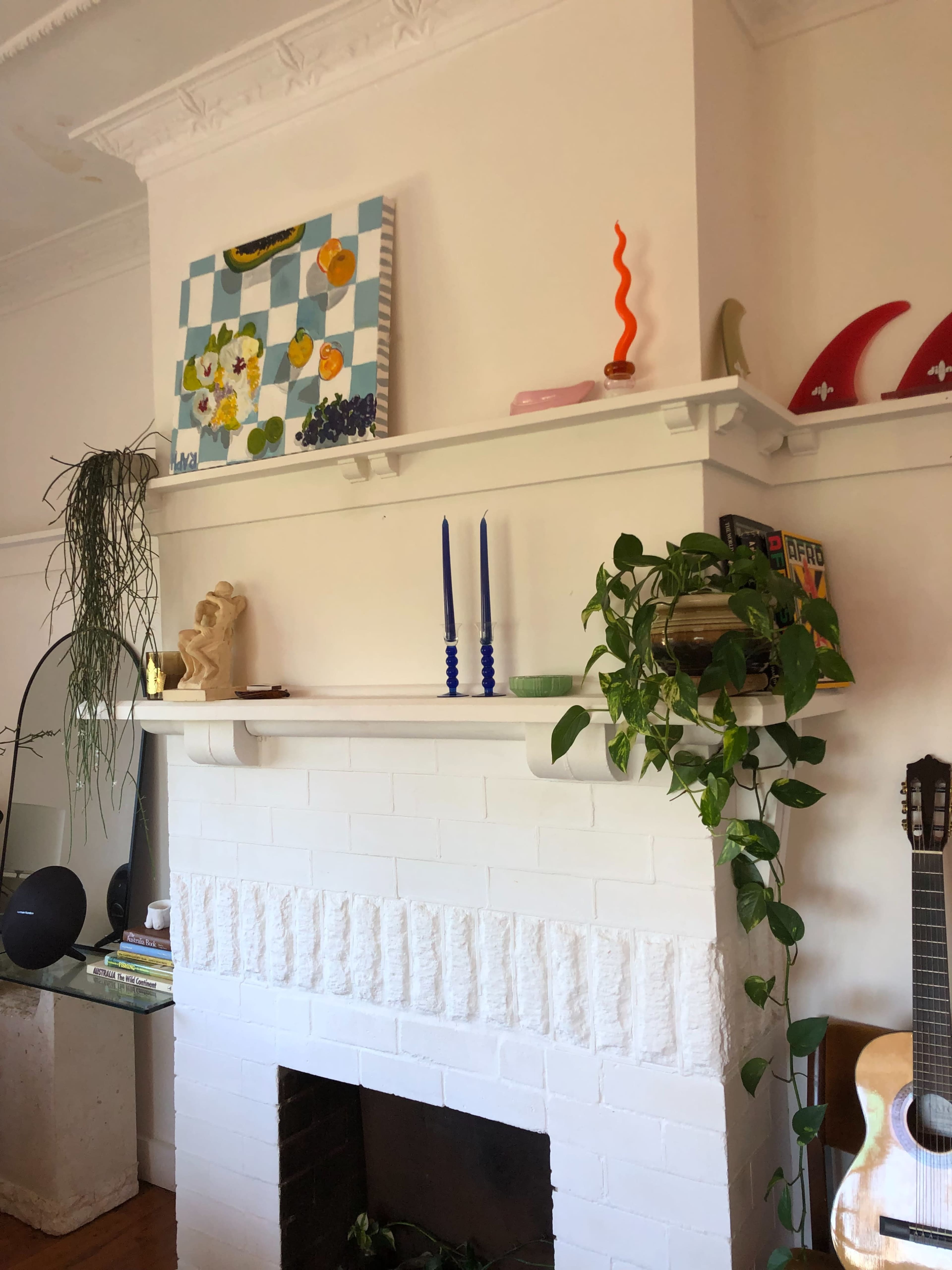 A white brick fireplace with a shelf displaying colorful decor items, candles, and plants, alongside a guitar leaning against the wall.