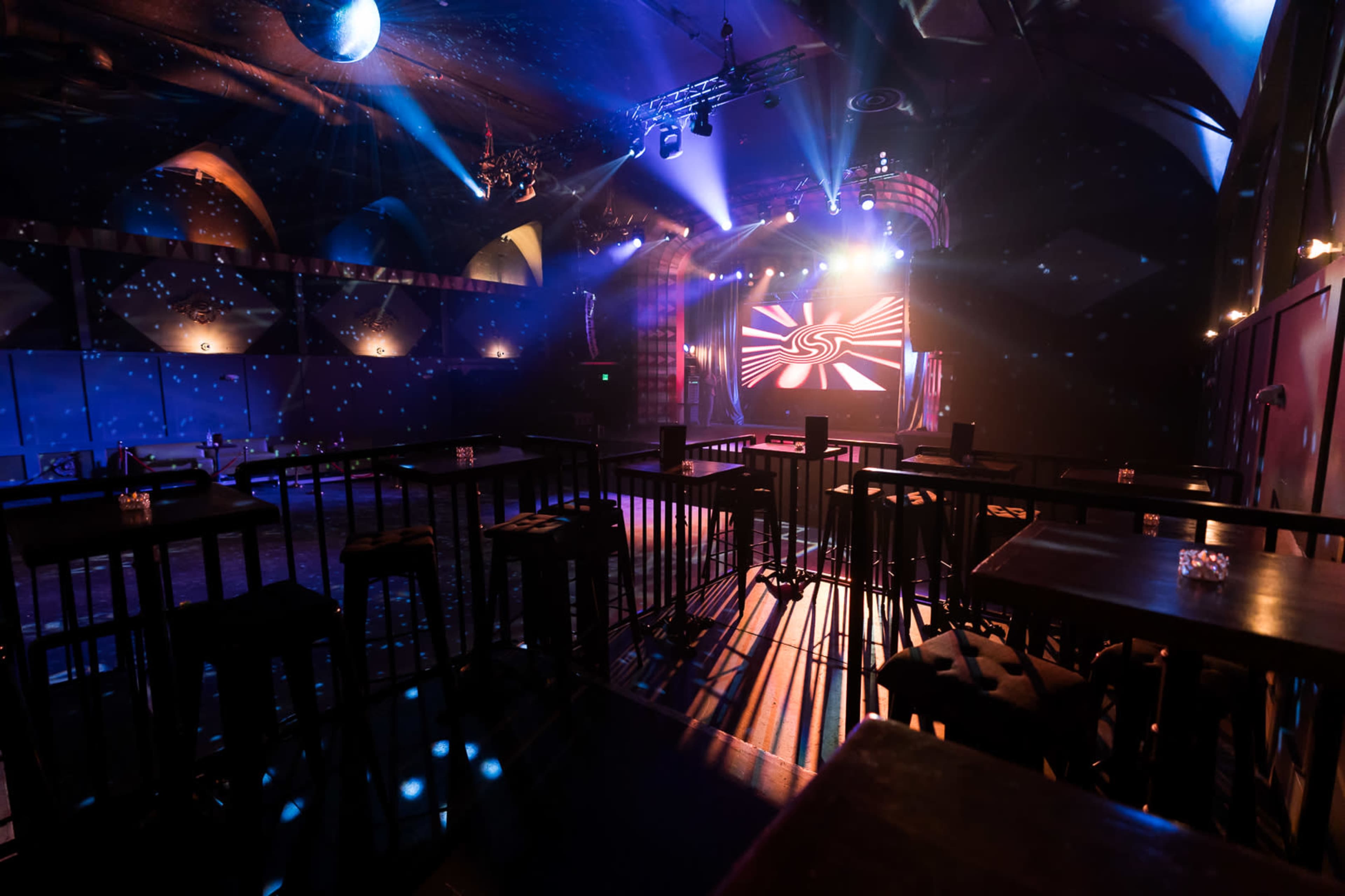 The image shows a dimly lit performance venue with seating arrangements, stage lighting, and a backdrop displaying a graphic design.