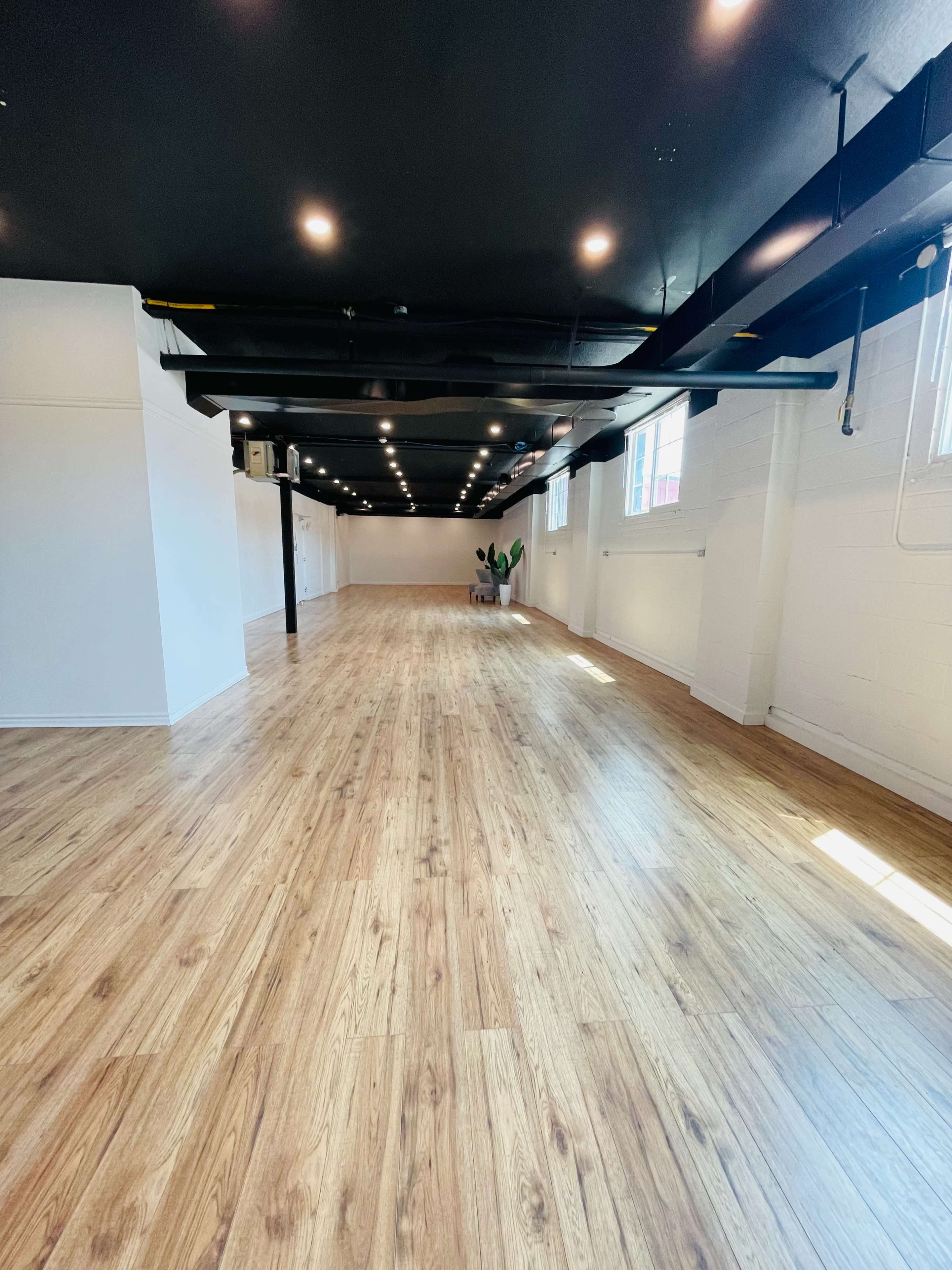 A spacious, empty room with wooden floors and black ceiling beams, featuring large windows that allow natural light to enter.