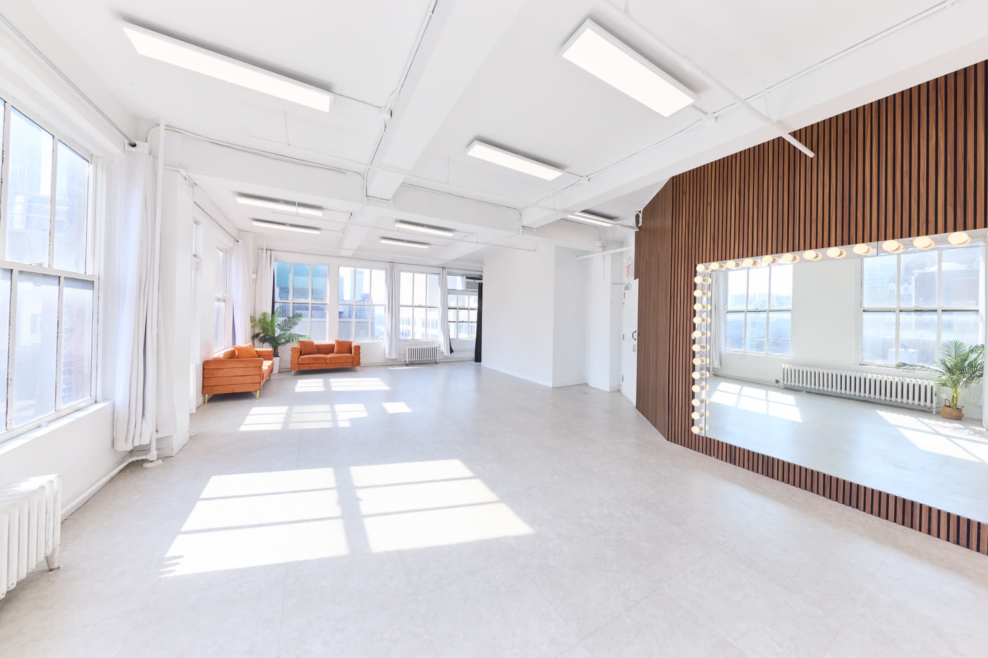 The image depicts a spacious, bright studio with large windows, a light-colored floor, an orange couch, and a mirror framed by light bulbs.