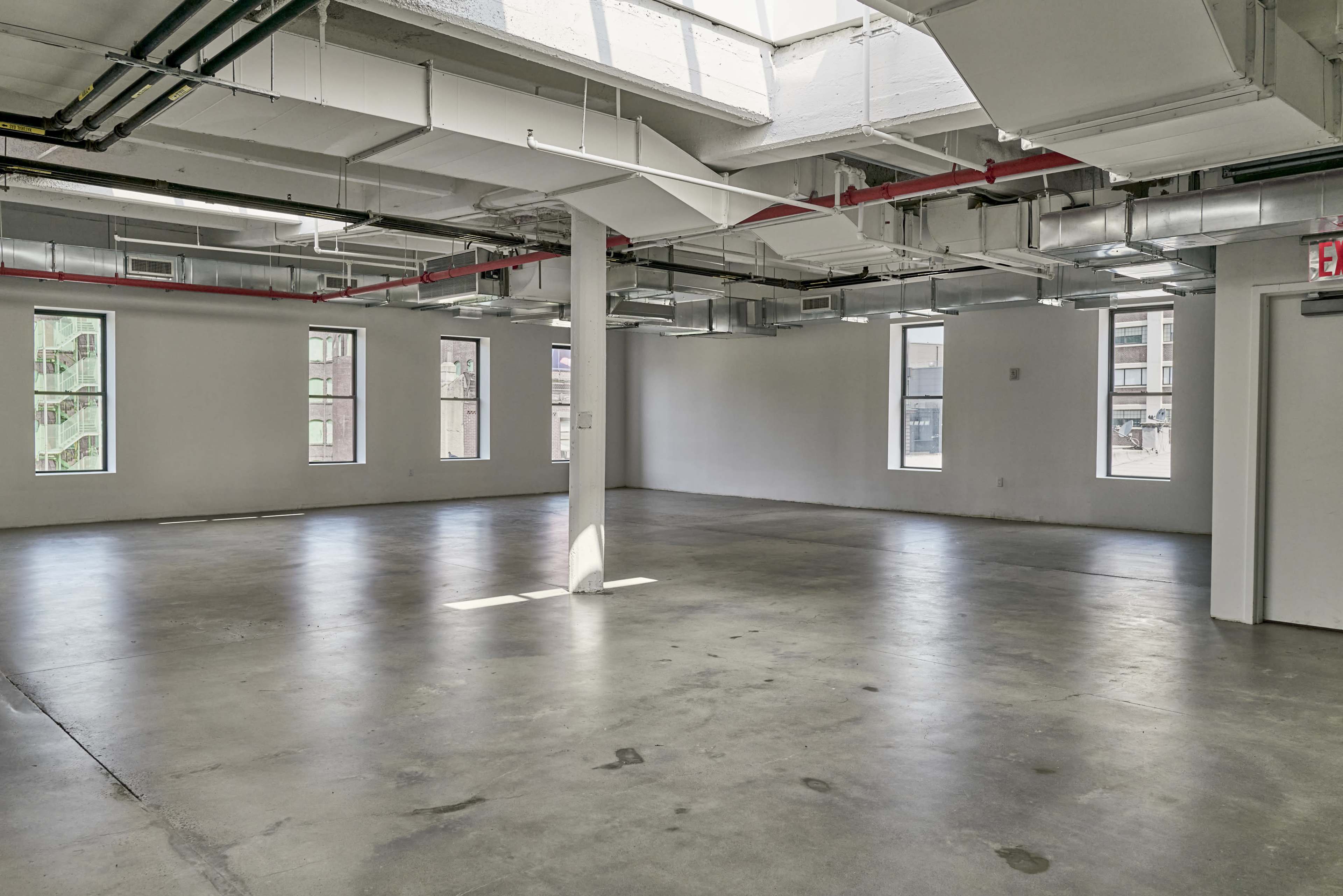 The image shows a spacious, empty room with concrete floors, large windows, and exposed ductwork.