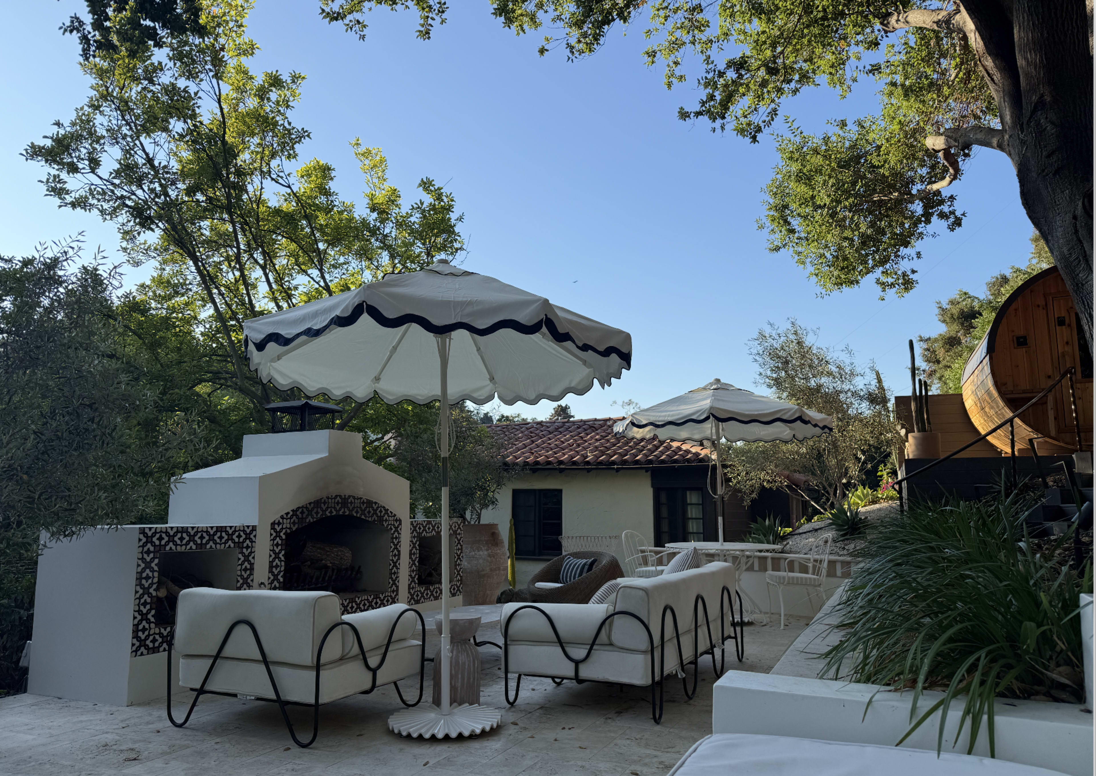 The scene features a patio area with a white seating arrangement, two large umbrellas, and a rustic outdoor fireplace, surrounded by trees and greenery.