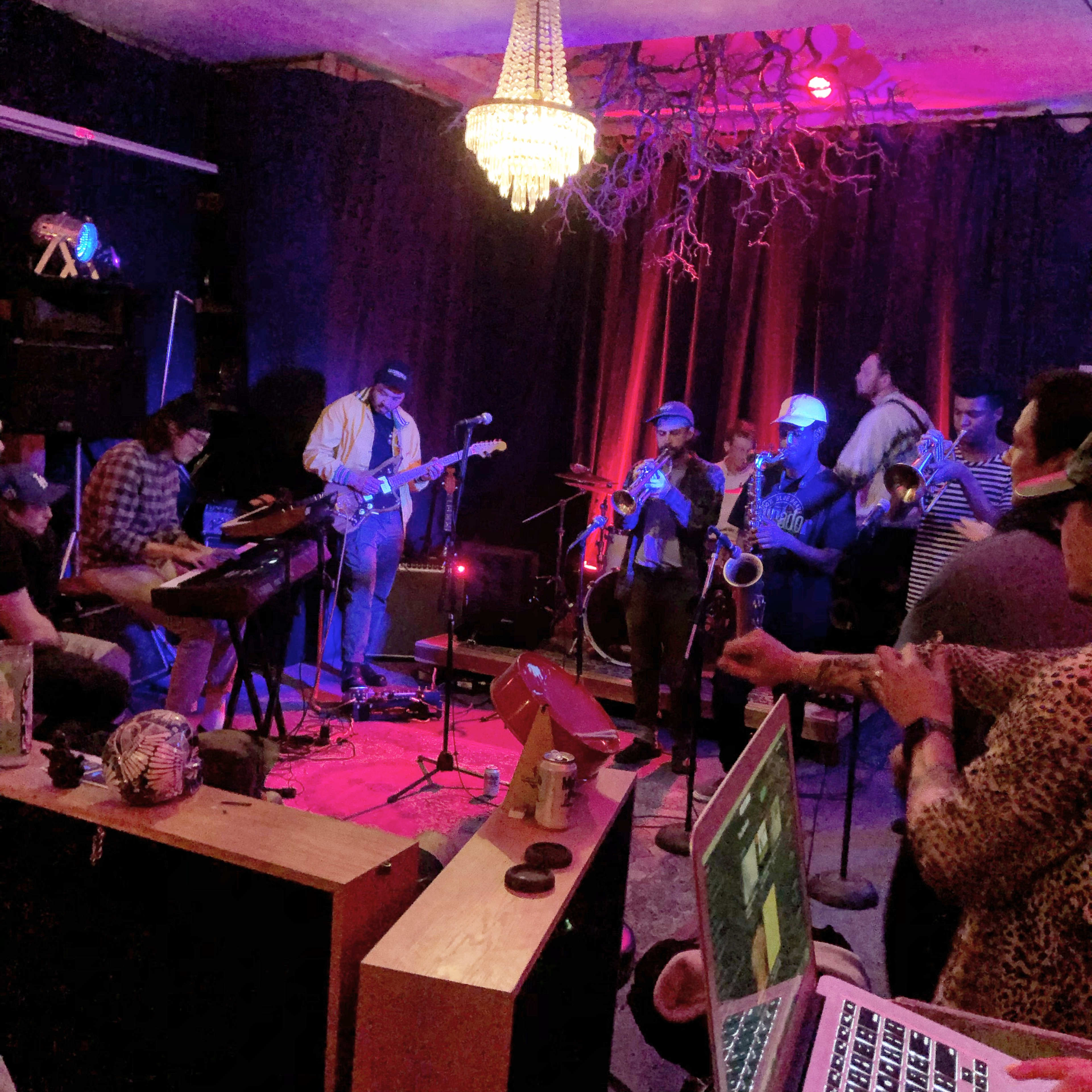 A live band performs on a small stage while an audience observes from a seating area in a dimly lit venue adorned with a chandelier.