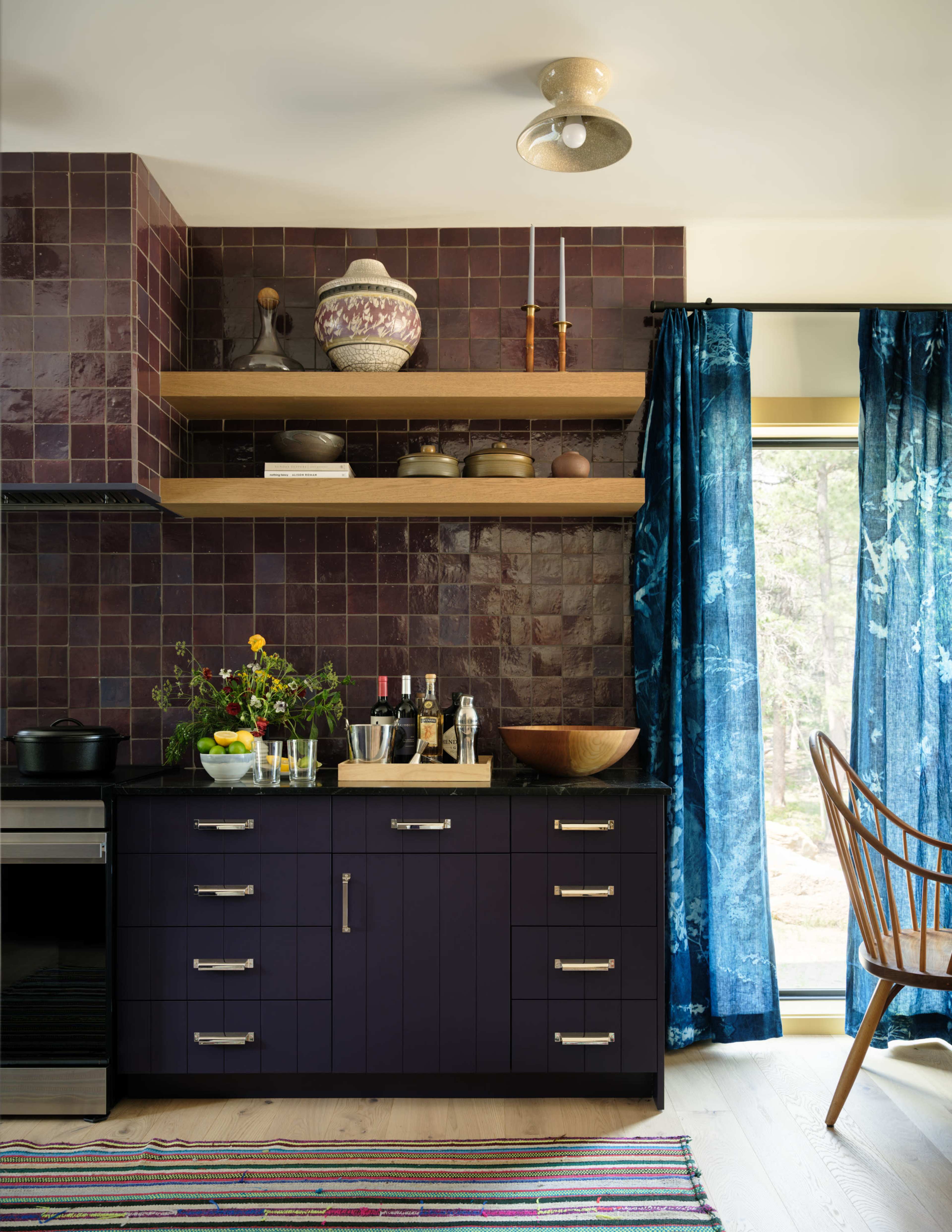 The kitchen features deep purple tiles, wooden shelves with various kitchen items, and a colorful rug under a window adorned with blue curtains.