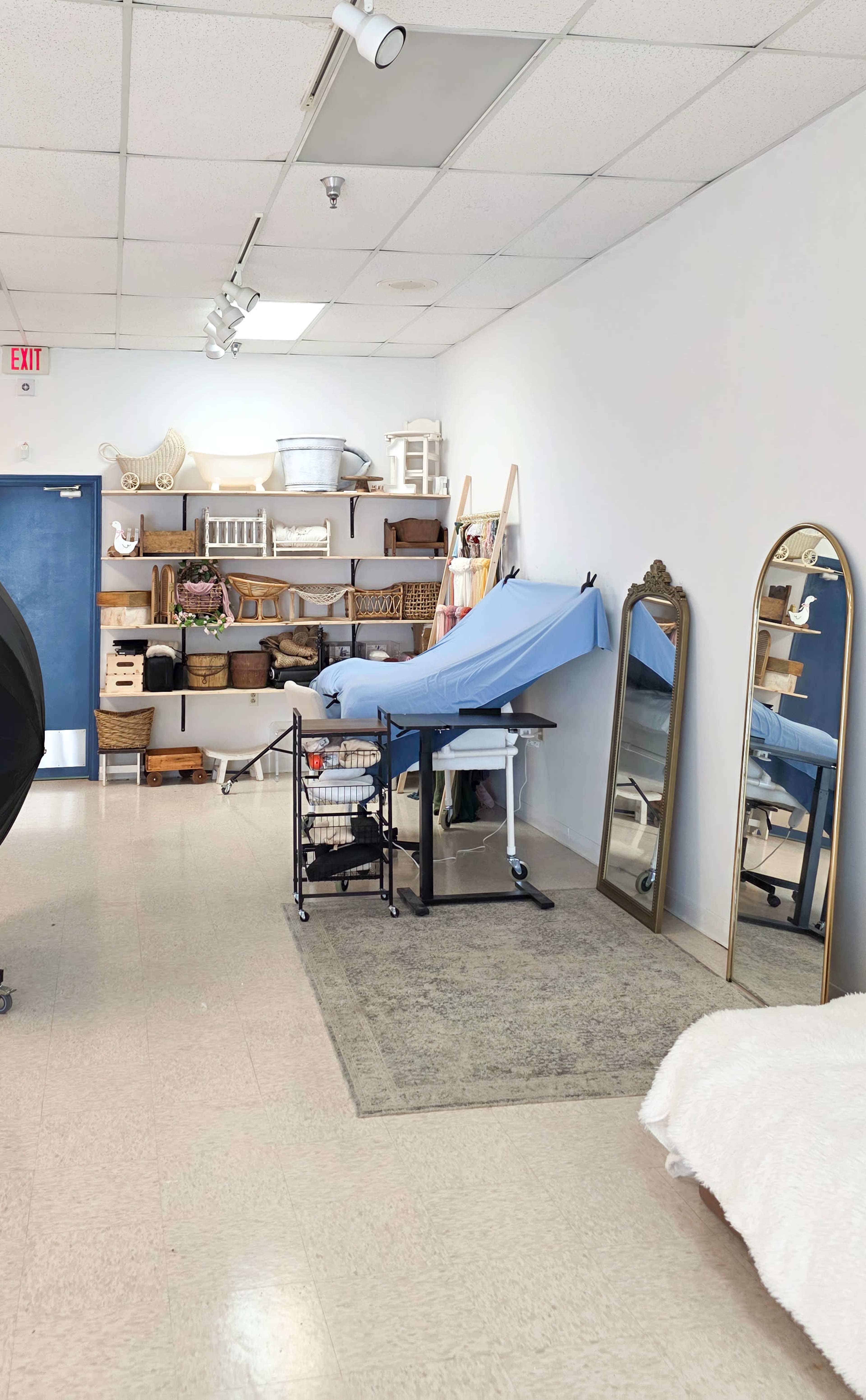 The image shows a neatly organized room with a treatment table covered in blue fabric, a large mirror, shelves with various storage baskets, and a roll cart in the foreground.