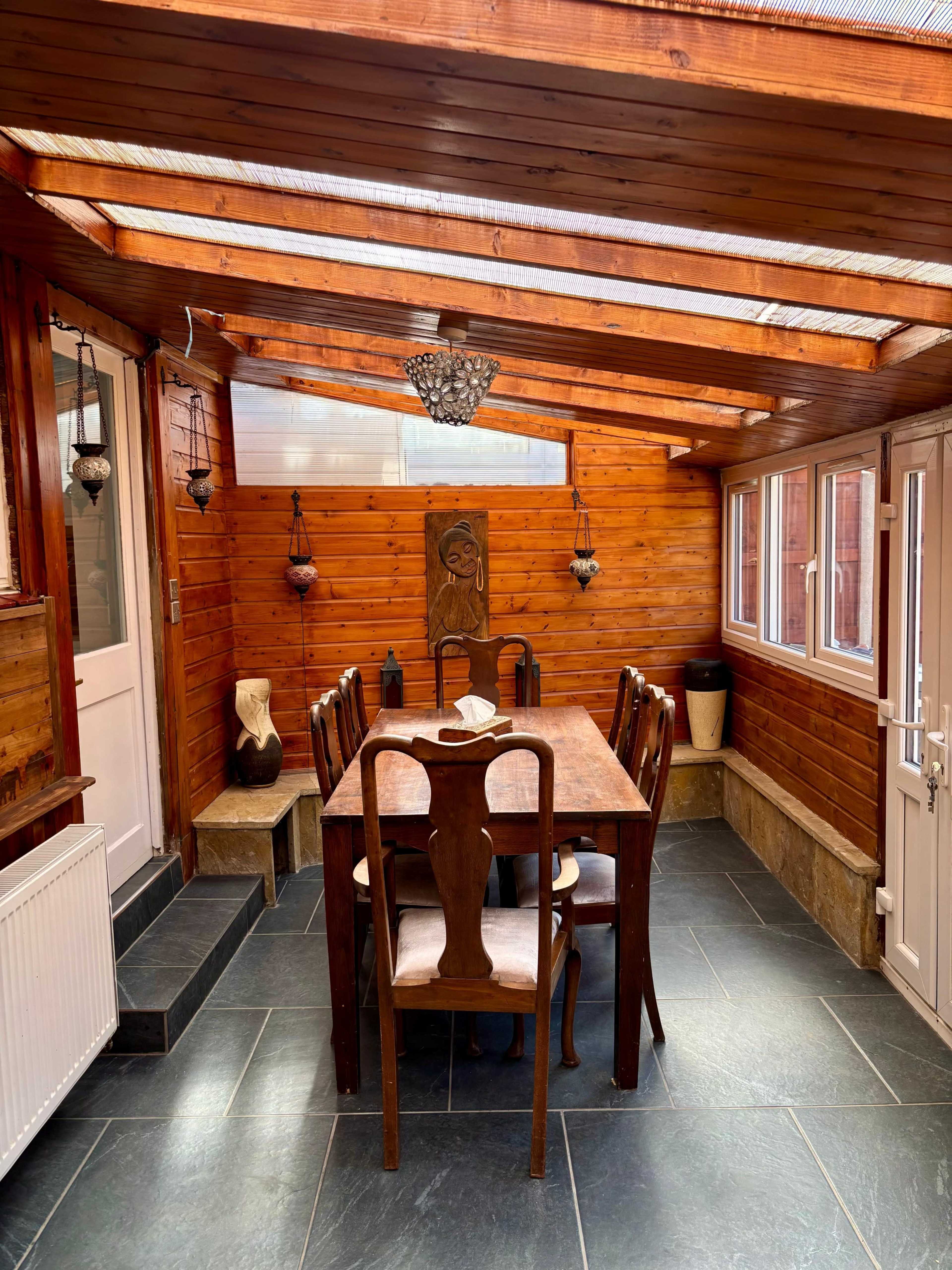The image shows a sunlit dining area with a wooden table and chairs, surrounded by wooden paneling and large windows.
