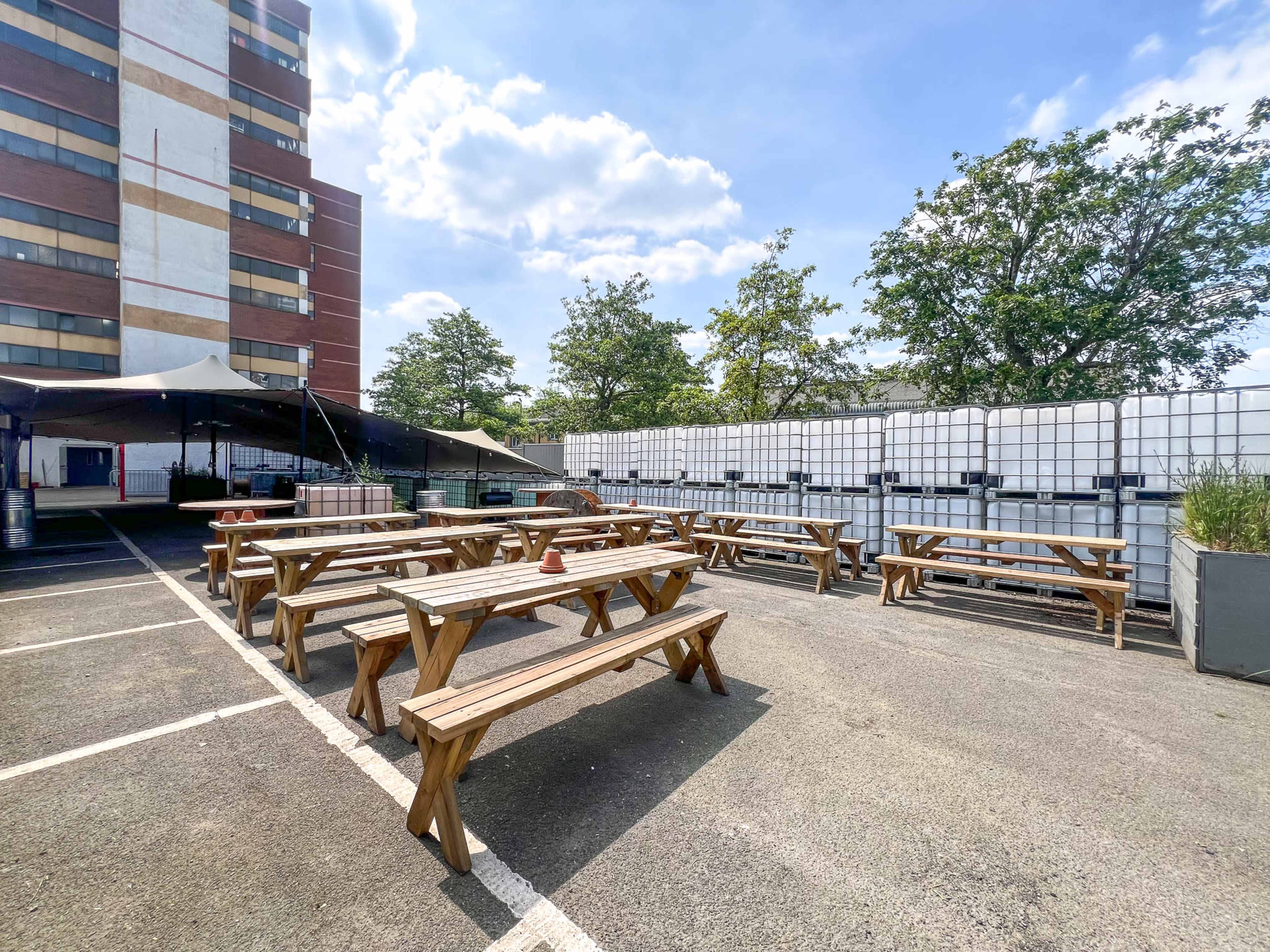 An outdoor area with several wooden picnic tables arranged neatly in a parking lot, bordered by large plastic water containers and a tall building in the background.