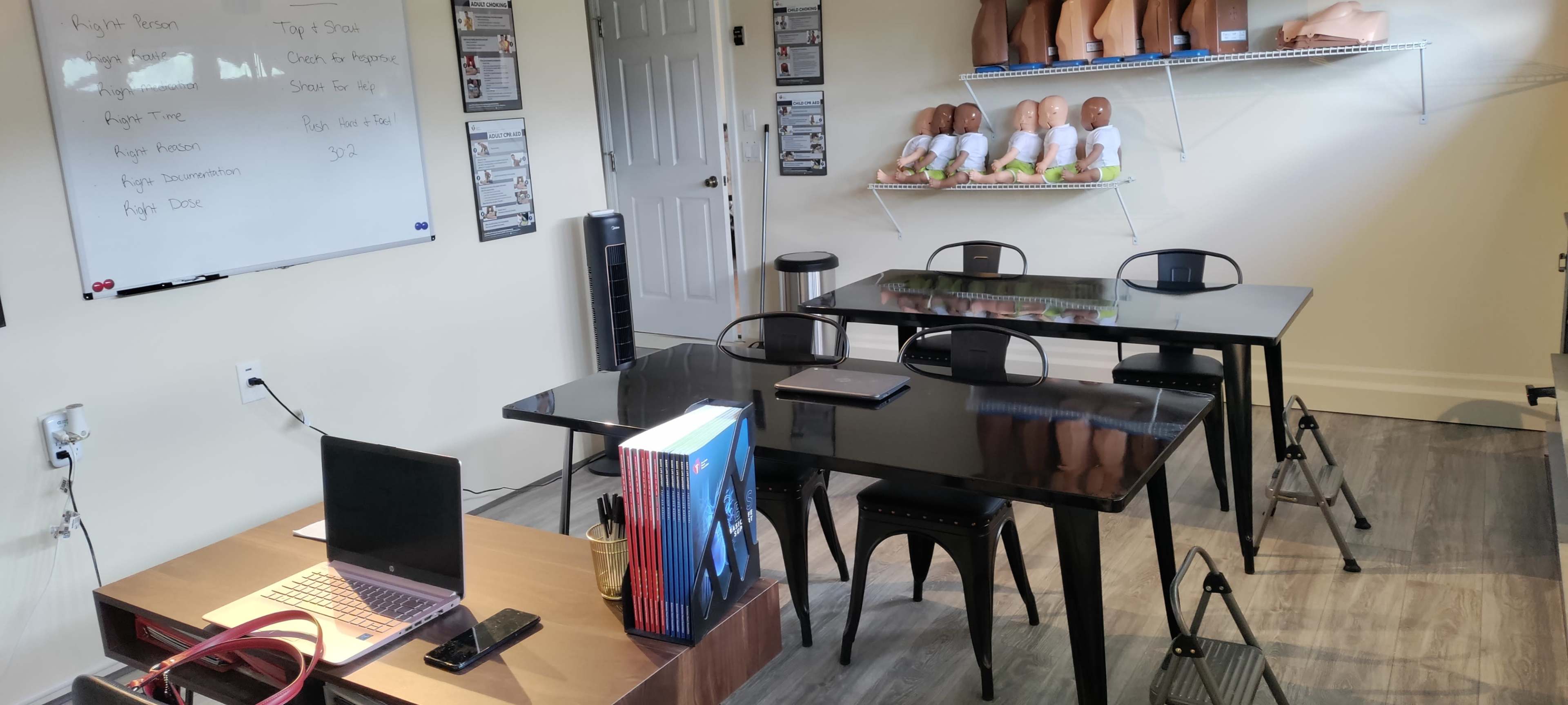 A training room with several black tables, chairs, a whiteboard, and shelves displaying mannequins and instructional materials.
