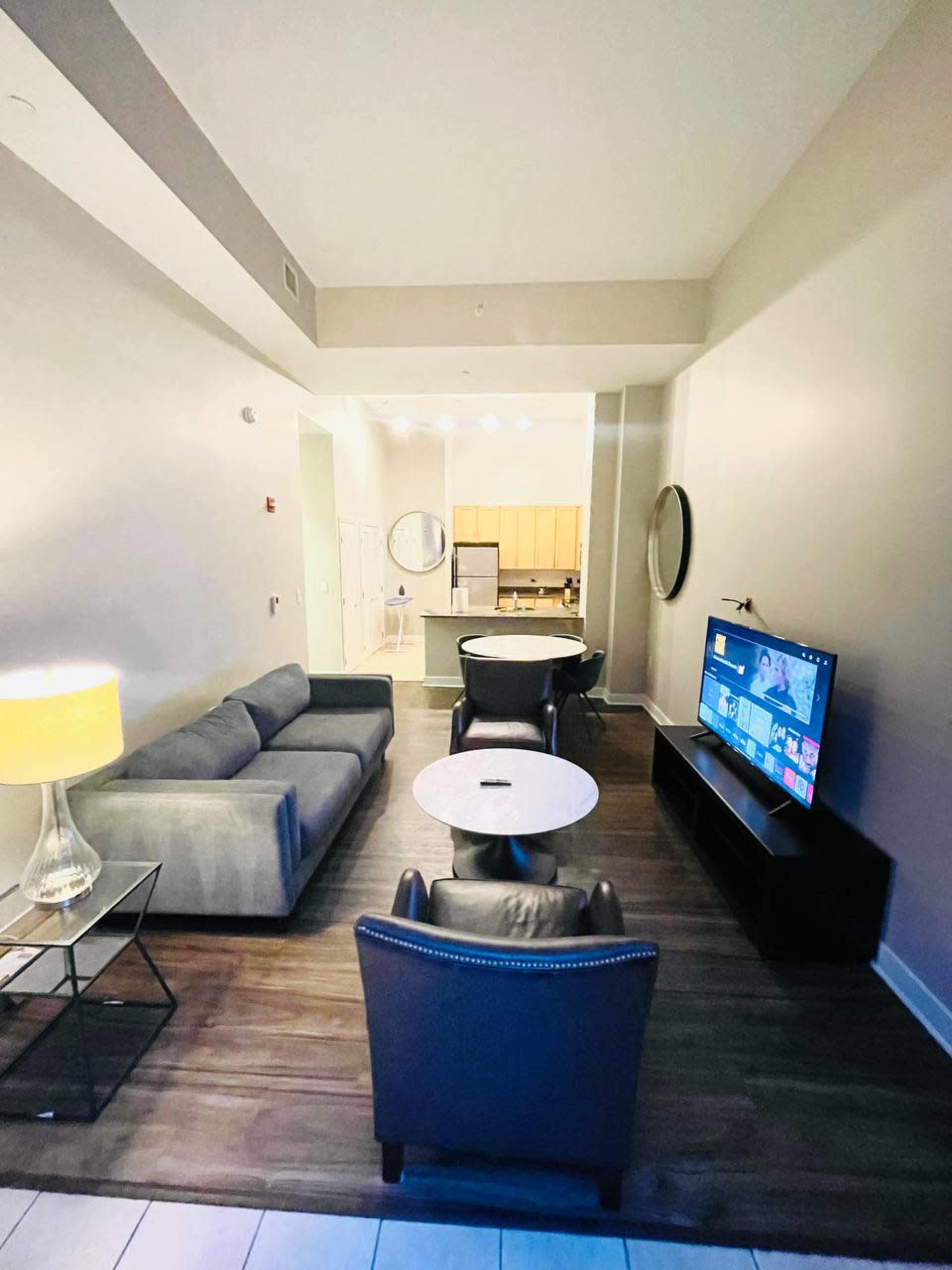 A modern living space features a gray couch, two chairs, a round coffee table, a television on a media console, and a view into a kitchen area.