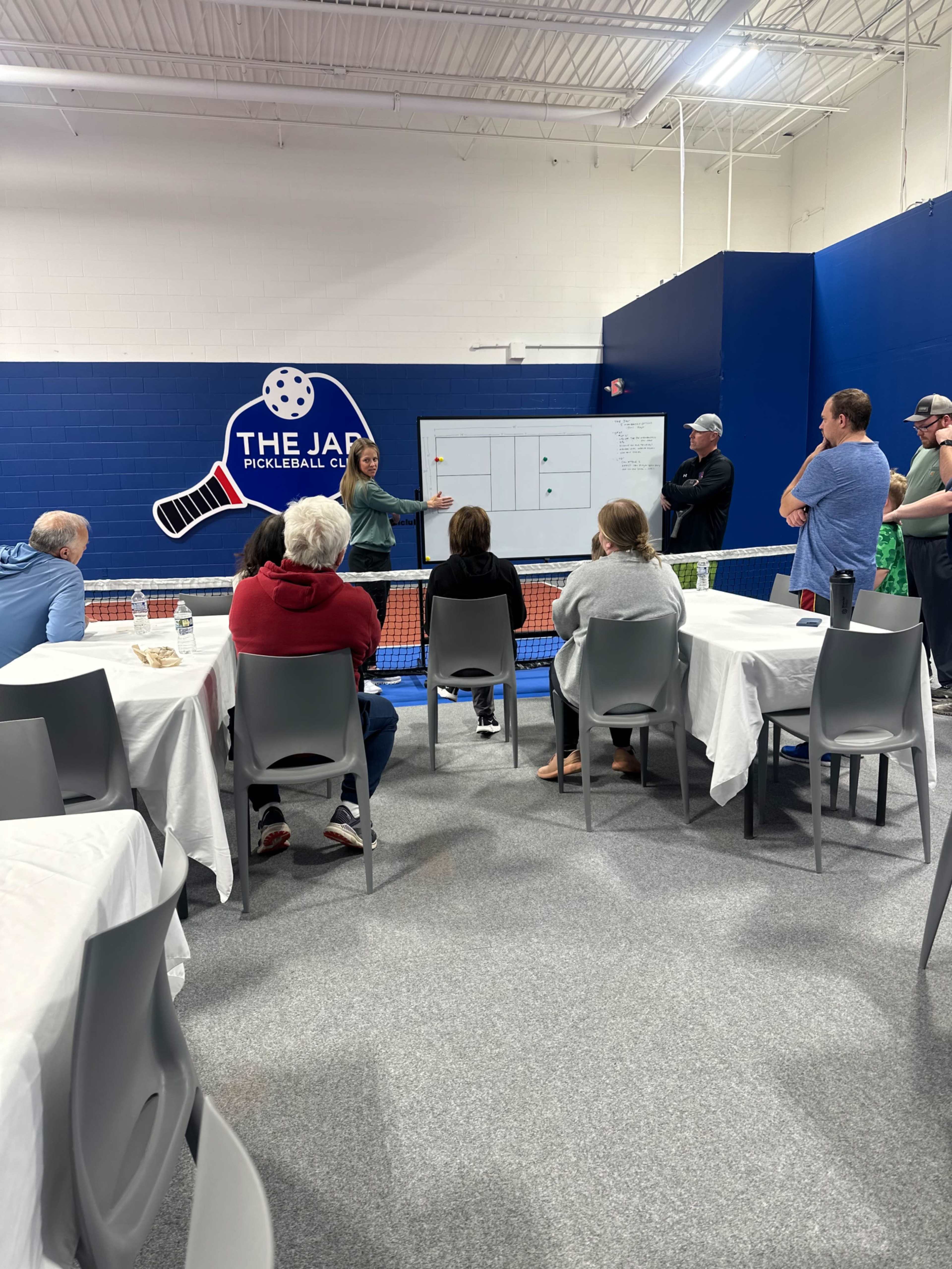 A group of people is gathered in a indoor pickleball facility, listening to a presentation near a pickleball court.