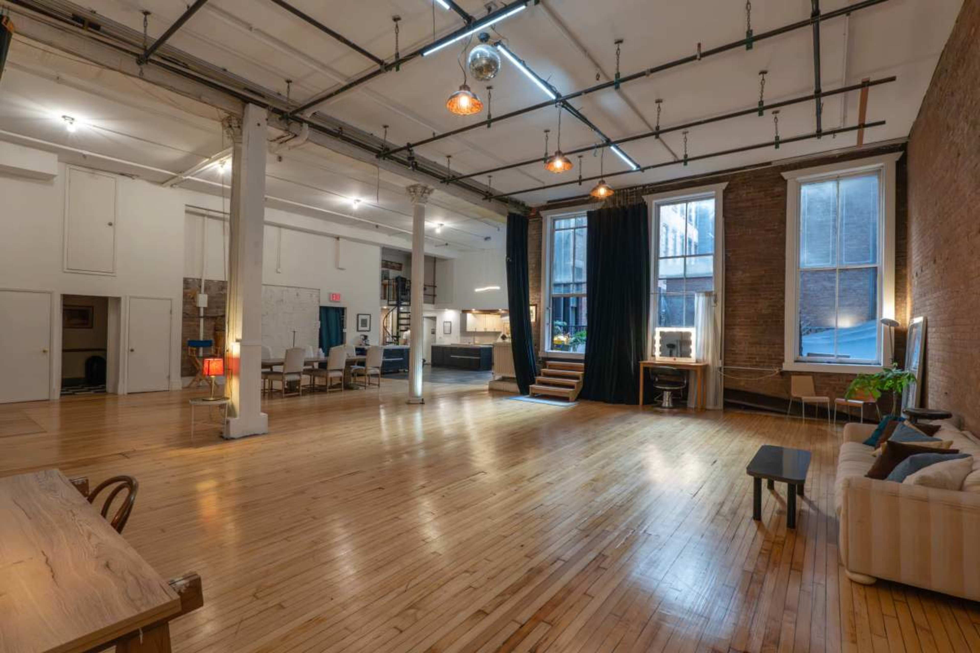 The image shows a spacious, multi-functional loft with wooden floors, large windows, and exposed brick walls, featuring a seating area, dining space, and kitchen visible in the background.