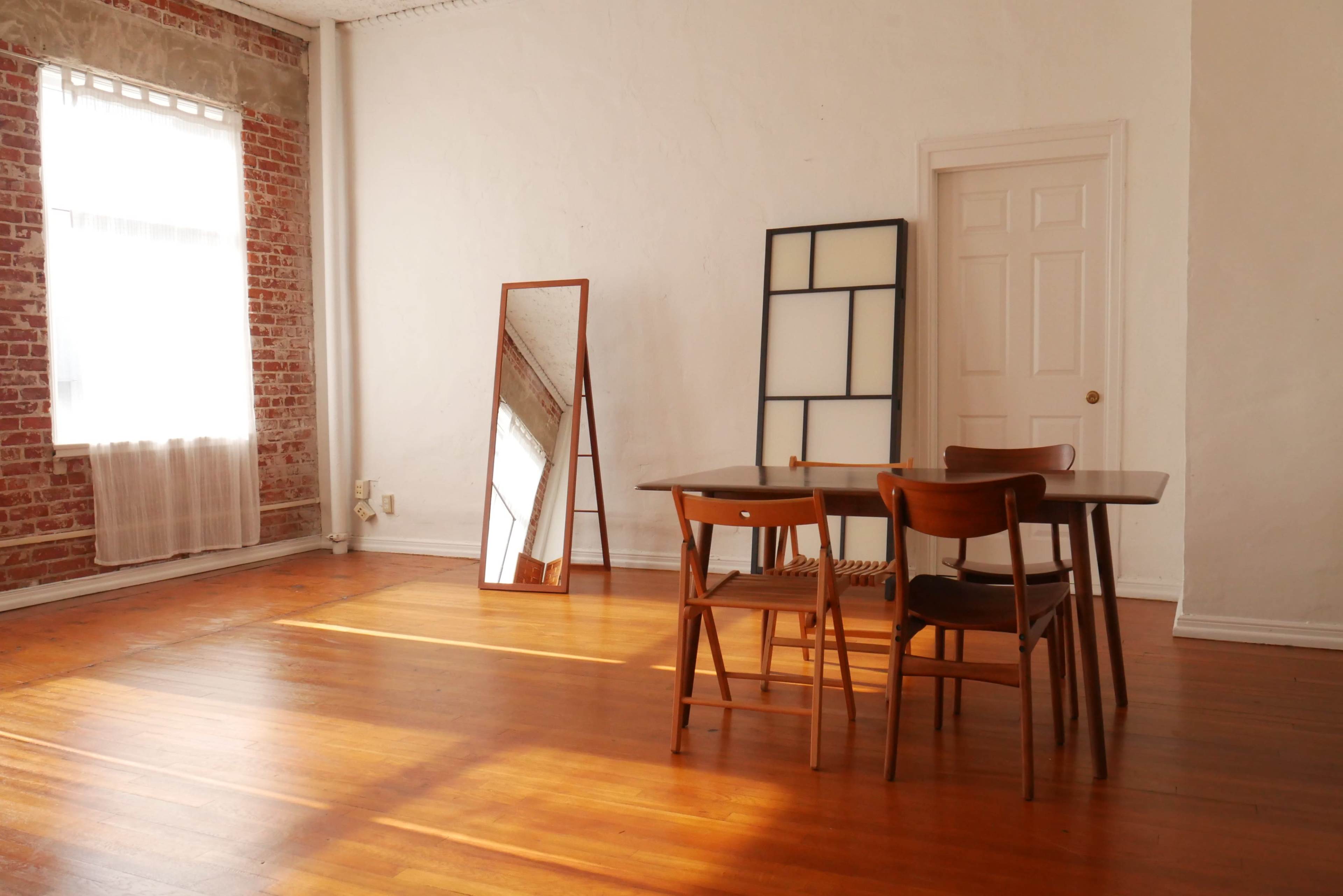 A well-lit room features a wooden table surrounded by chairs, a large mirror, and a door, with exposed brick walls and wooden flooring.