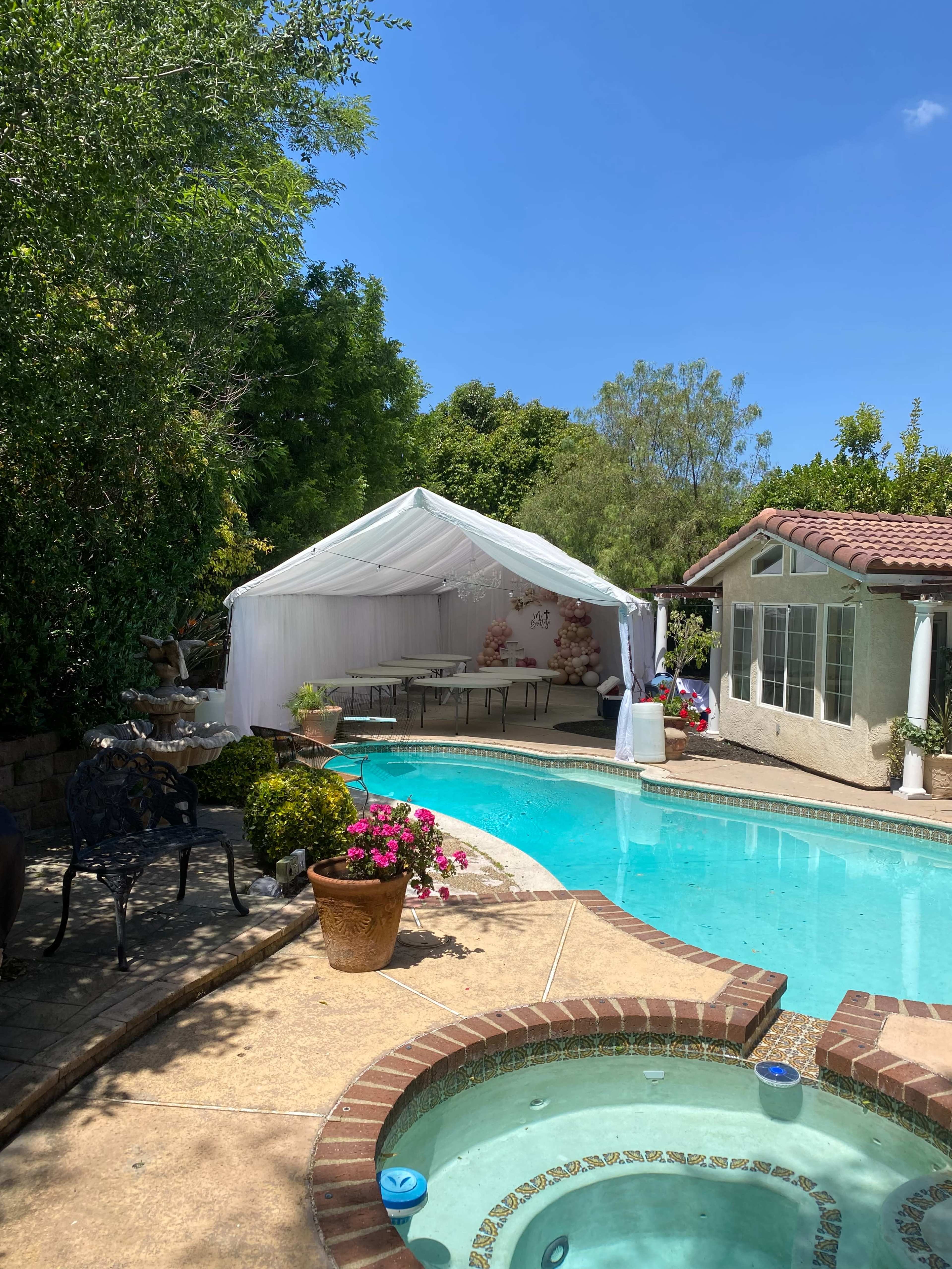 A white tent is setup beside a swimming pool in a backyard surrounded by greenery.