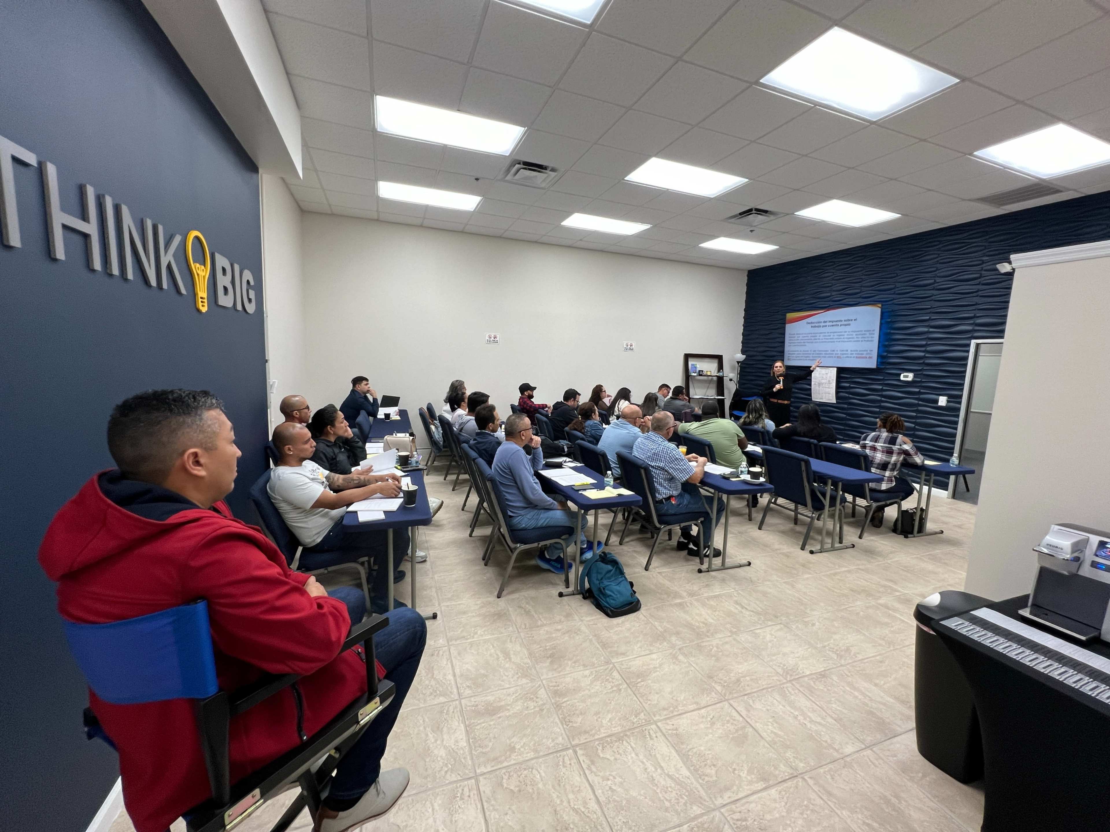 A group of individuals is seated at tables in a training room, listening to a speaker presenting in front of a large screen.