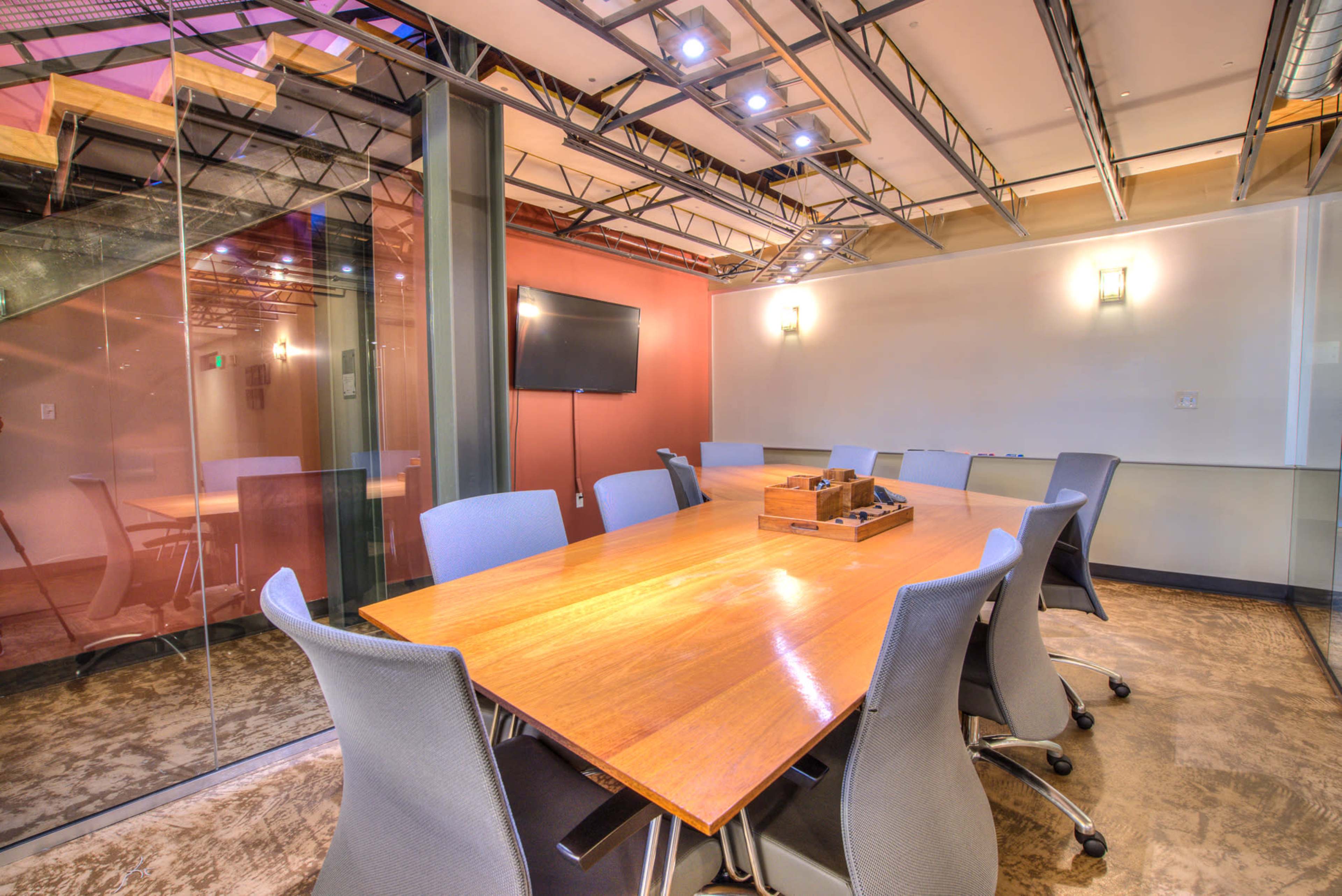 A conference room features a large wooden table surrounded by gray chairs, with a TV screen mounted on the wall and glass partitioning.
