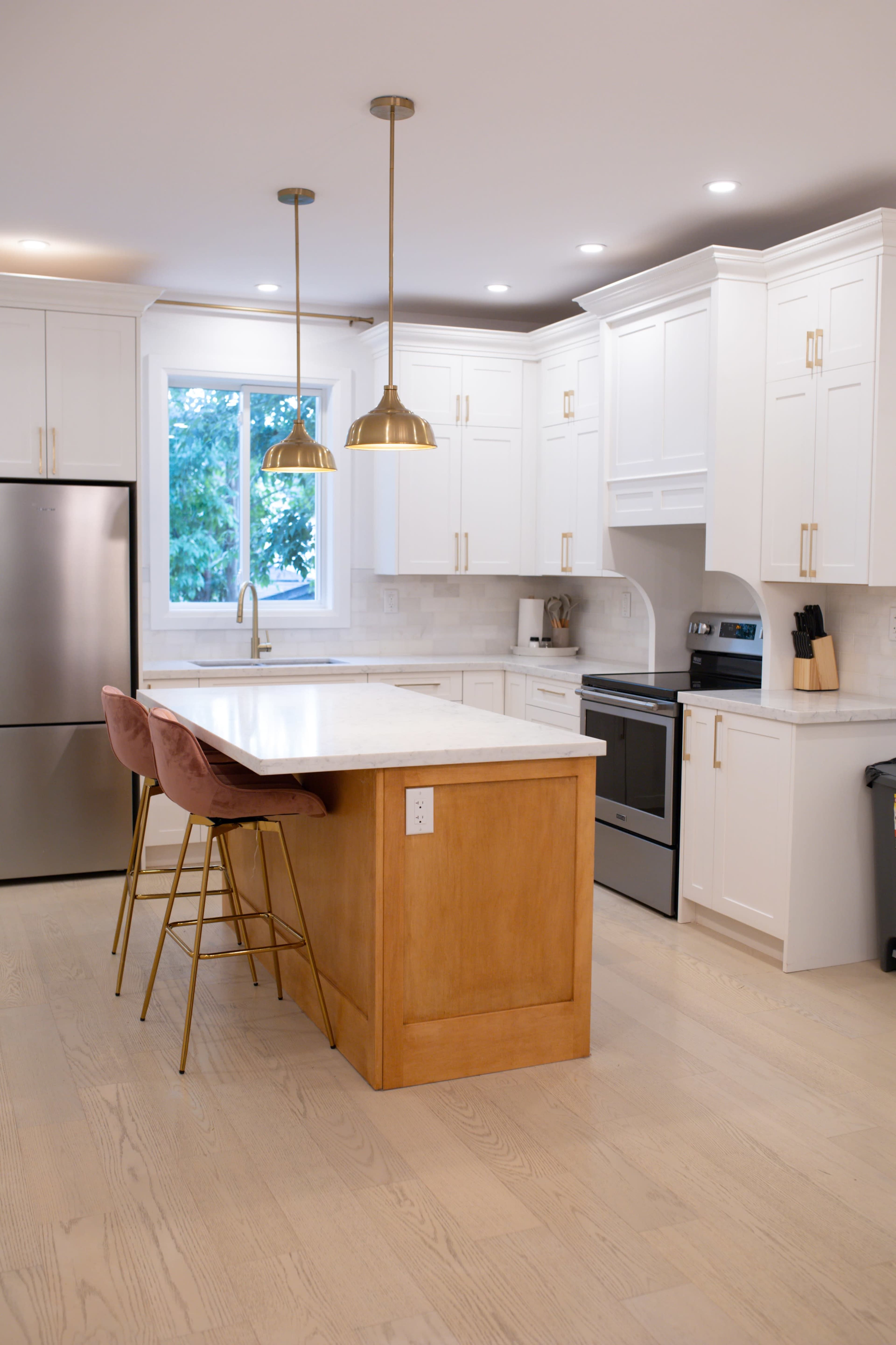 A modern kitchen features white cabinetry, stainless steel appliances, a large island with a wooden base, and two pink bar stools under gold pendant lights.