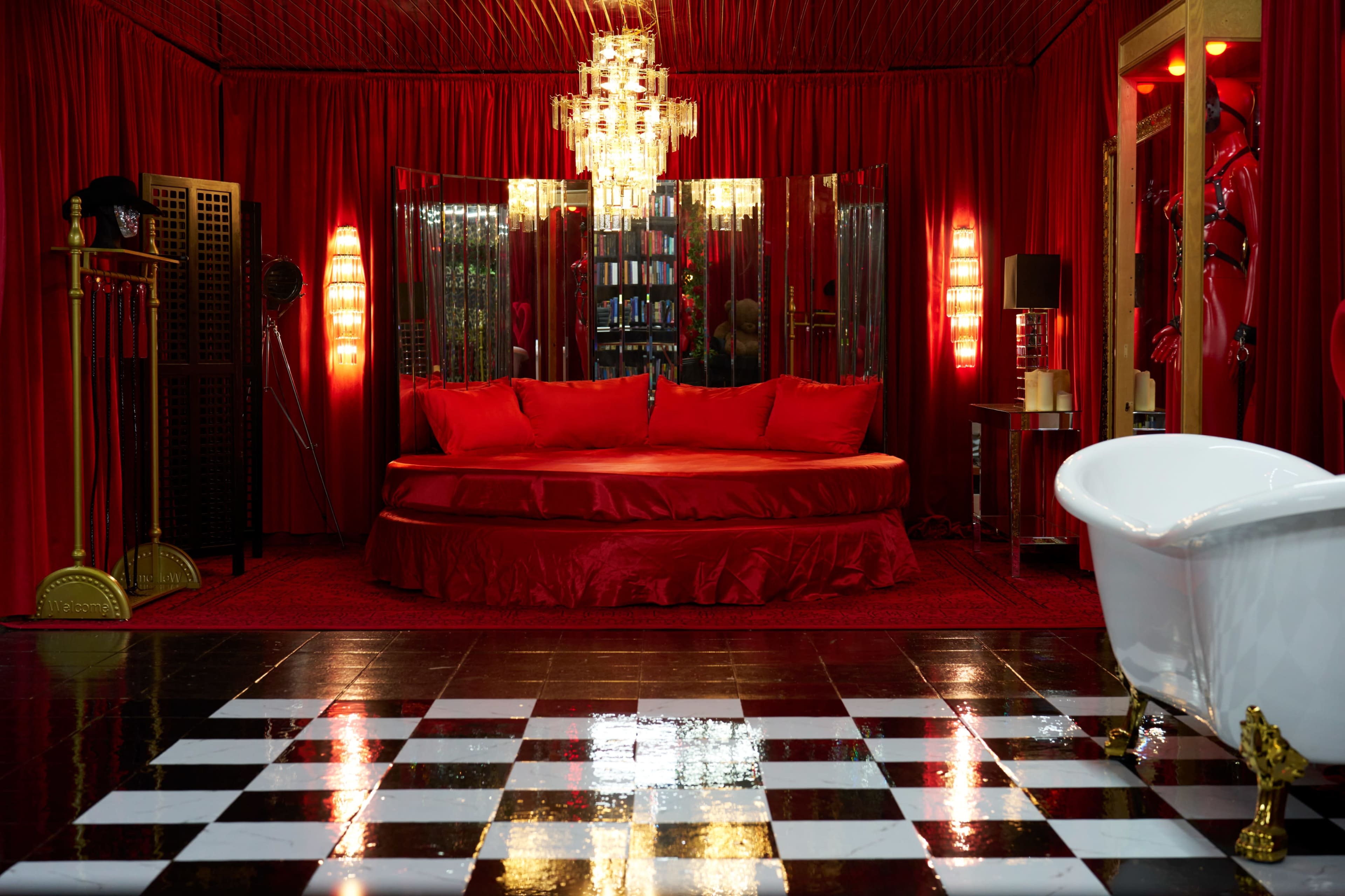 The image shows a lavish room decorated in red with a round bed at the center, surrounded by mirrored walls and a contrasting black and white checkered floor.