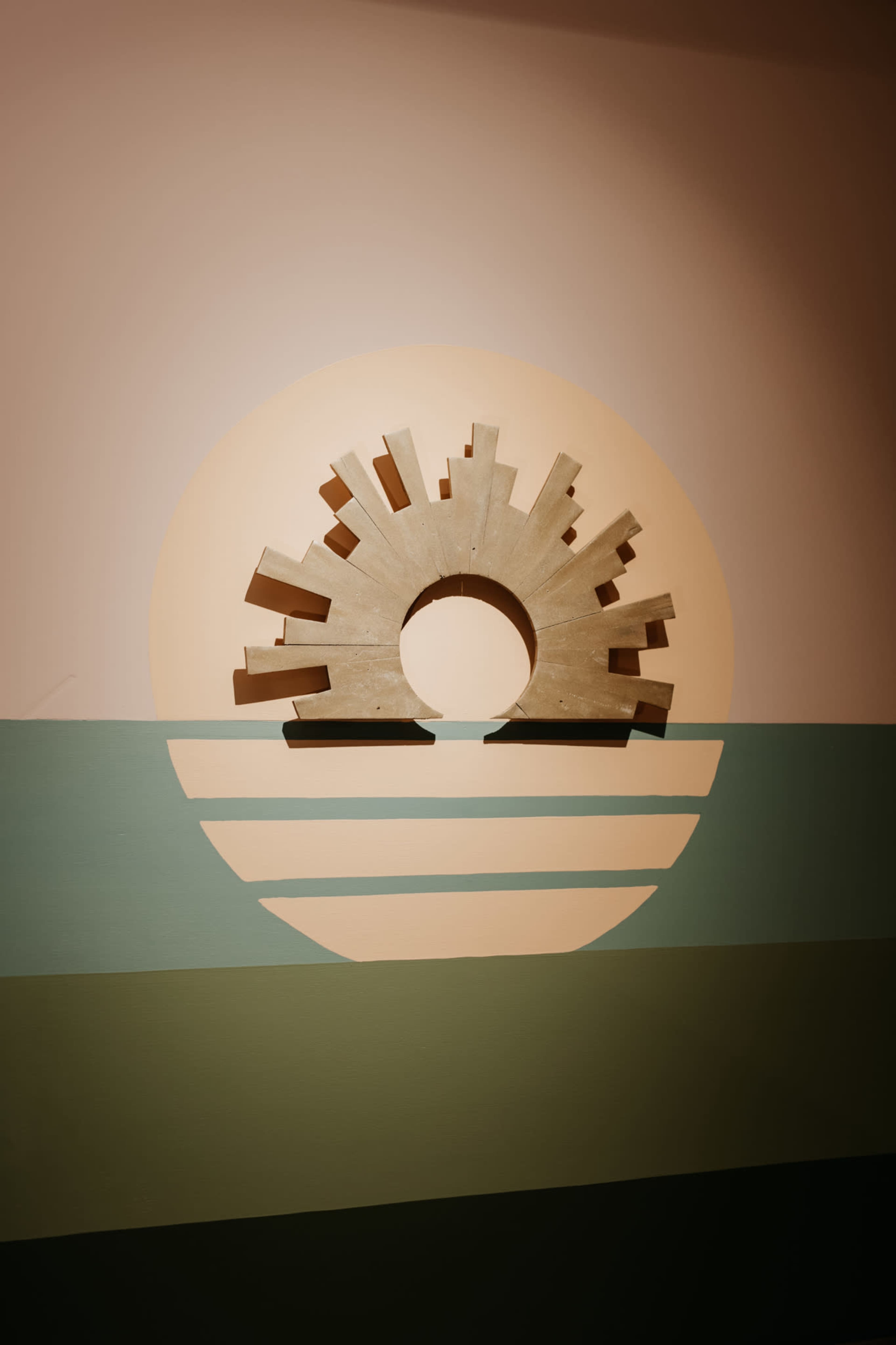 A circular sculpture resembling a sun rises above stylized horizontal bands of color on a wall.