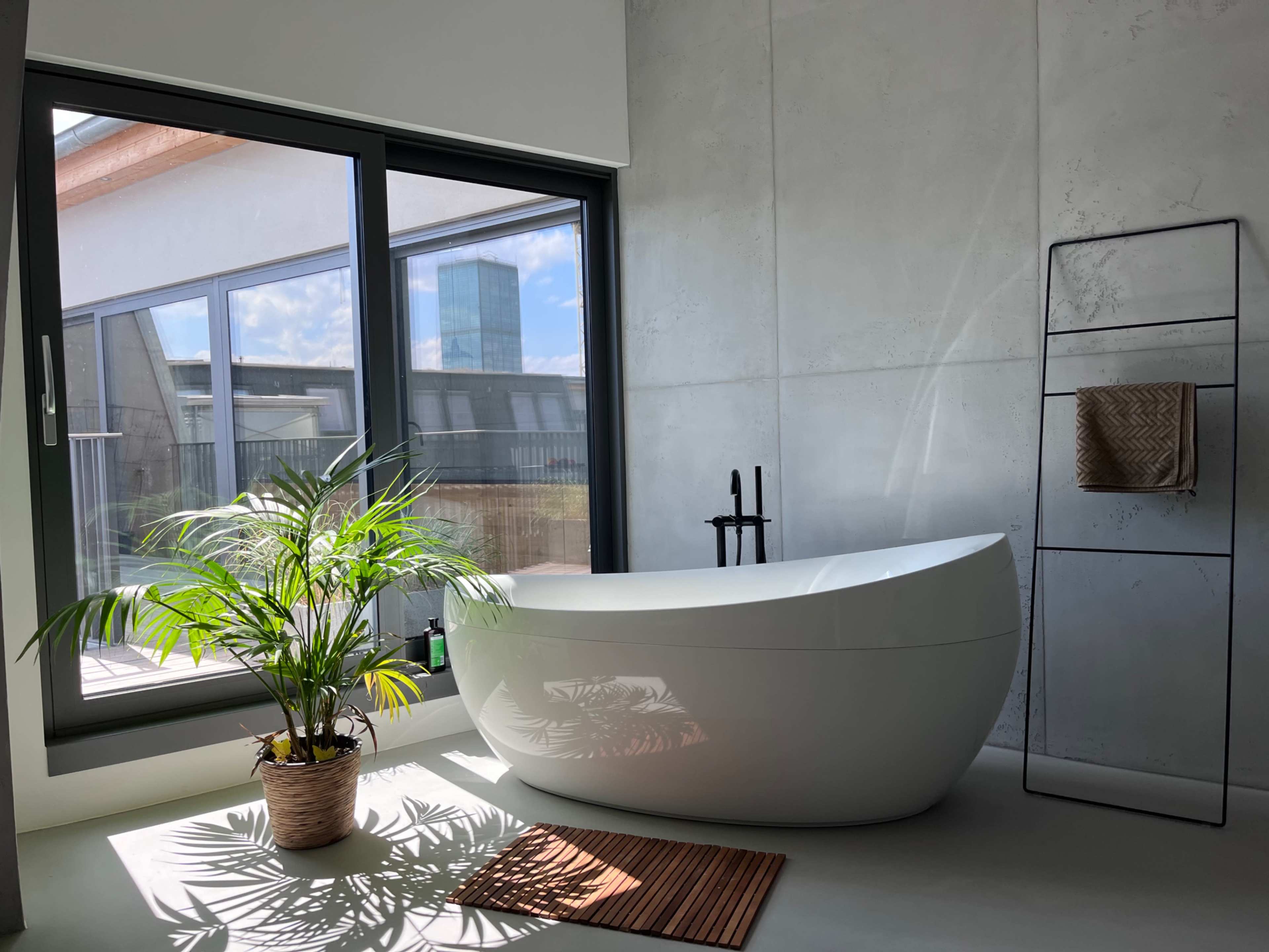 The image depicts a modern bathroom featuring a freestanding bathtub beside a large window with a view, complemented by a potted plant and a bath mat on the floor.
