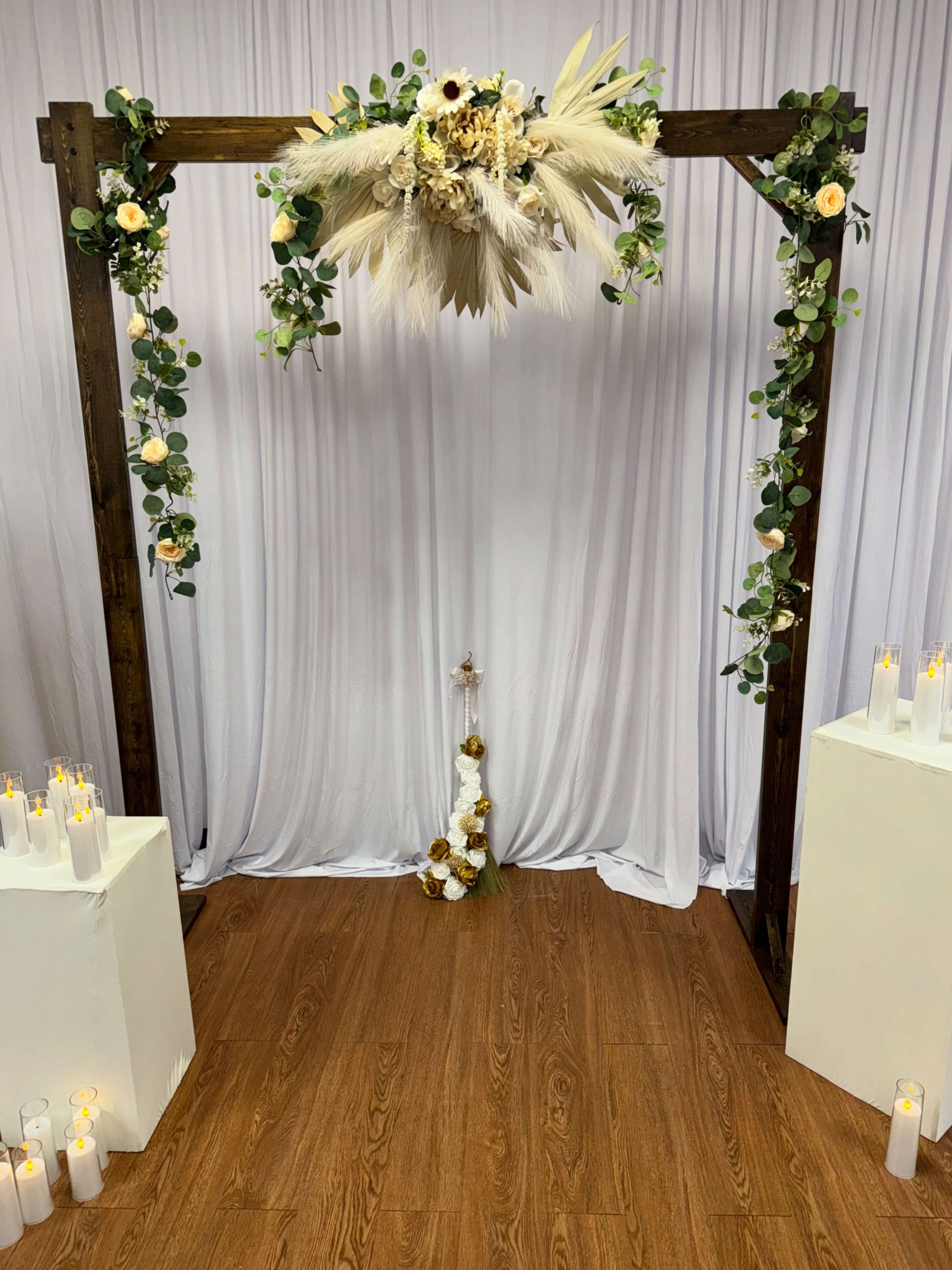 A wooden arch adorned with floral arrangements and greenery is set against a white backdrop, flanked by white pedestals and candles on the floor.