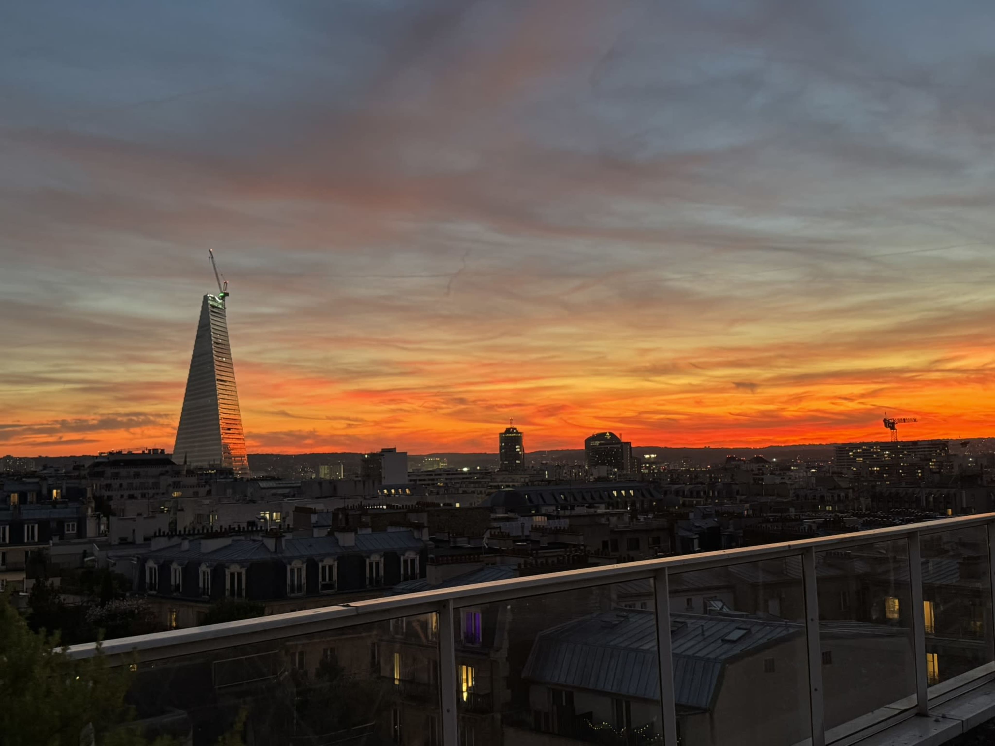 The image shows a skyline view of Paris at sunset, featuring the distinctive modern architecture of a tall building illuminated against a vibrant orange and pink sky.