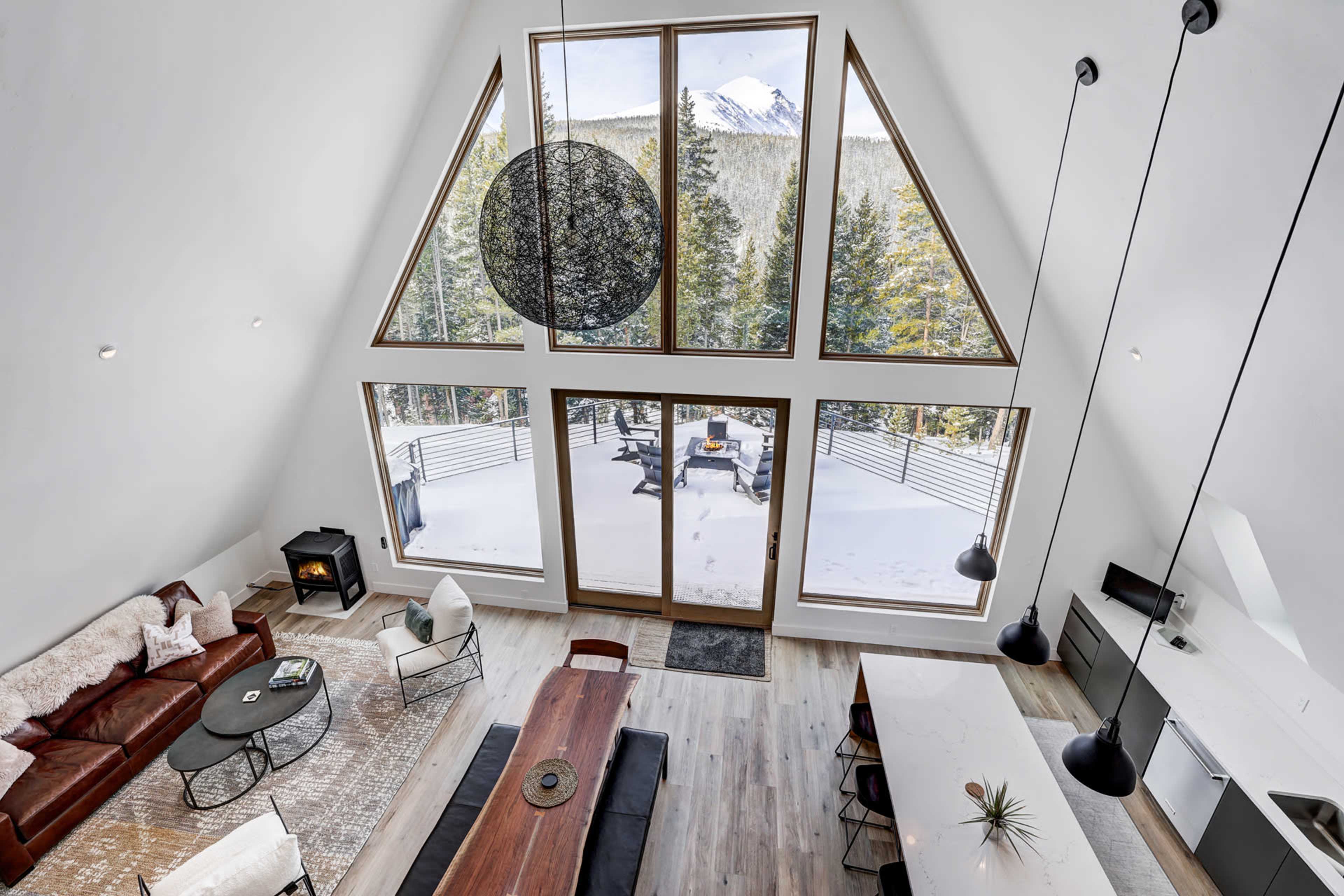 A modern A-frame cabin interior with large windows overlooking a snowy landscape, featuring a wooden dining table, a cozy seating area, and a fire stove.