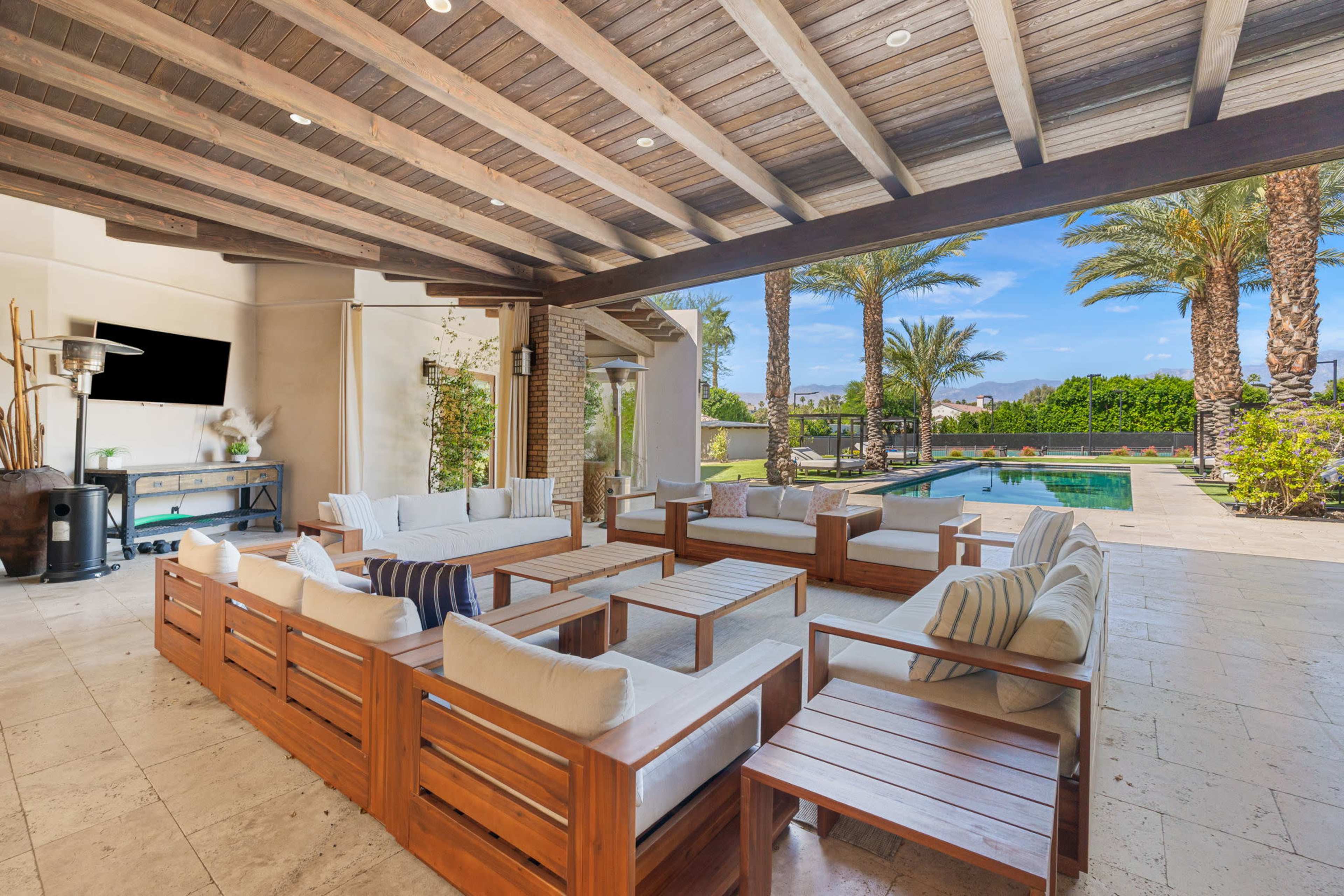 The image shows a shaded outdoor living area with wooden furniture and a pool visible in the background surrounded by palm trees.