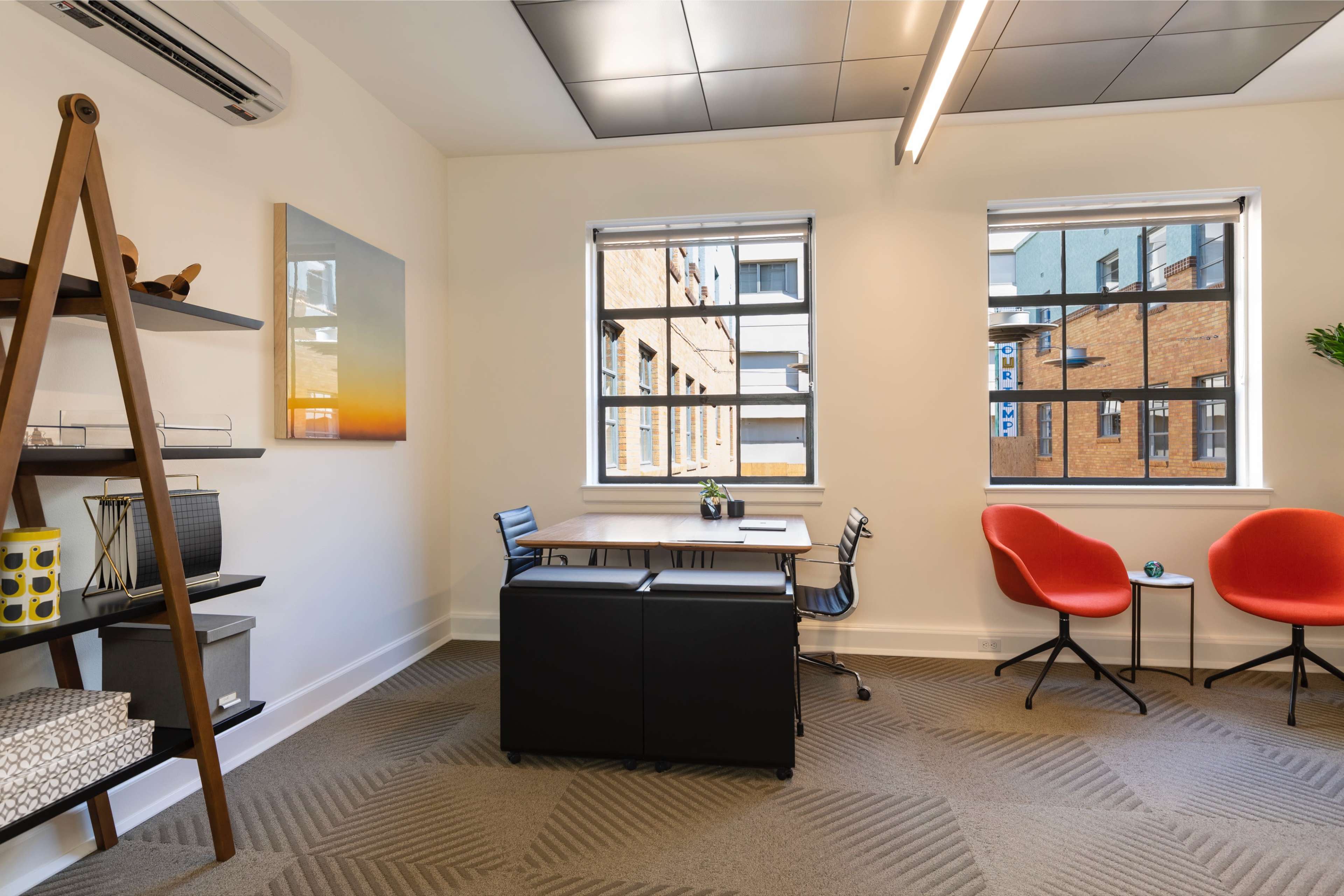 The image shows a modern office space with a desk and chair, two red chairs, a shelf, and windows overlooking a brick building.