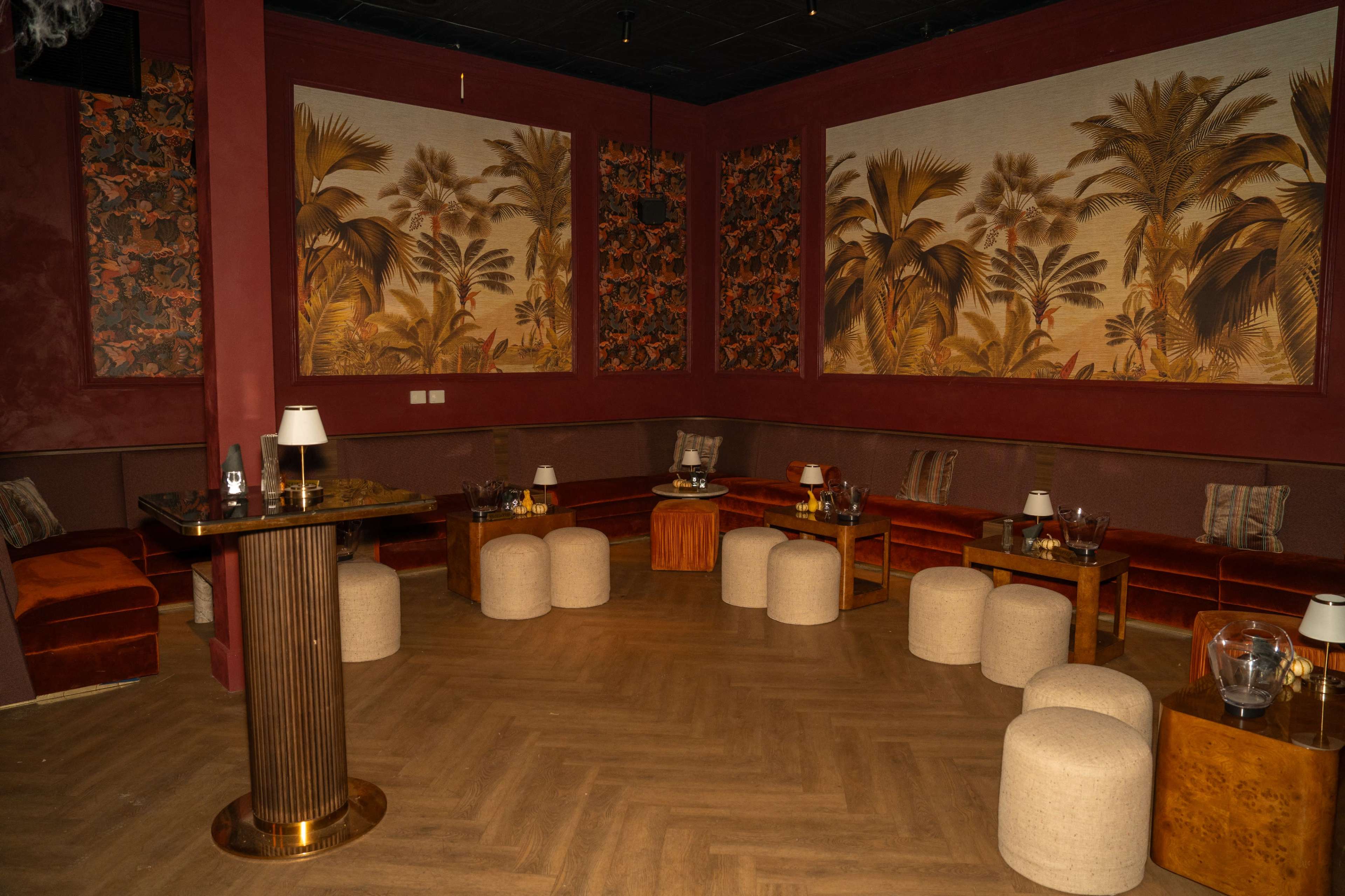 The image depicts a dimly lit lounge featuring red walls adorned with tropical-themed murals, plush seating, and small tables.