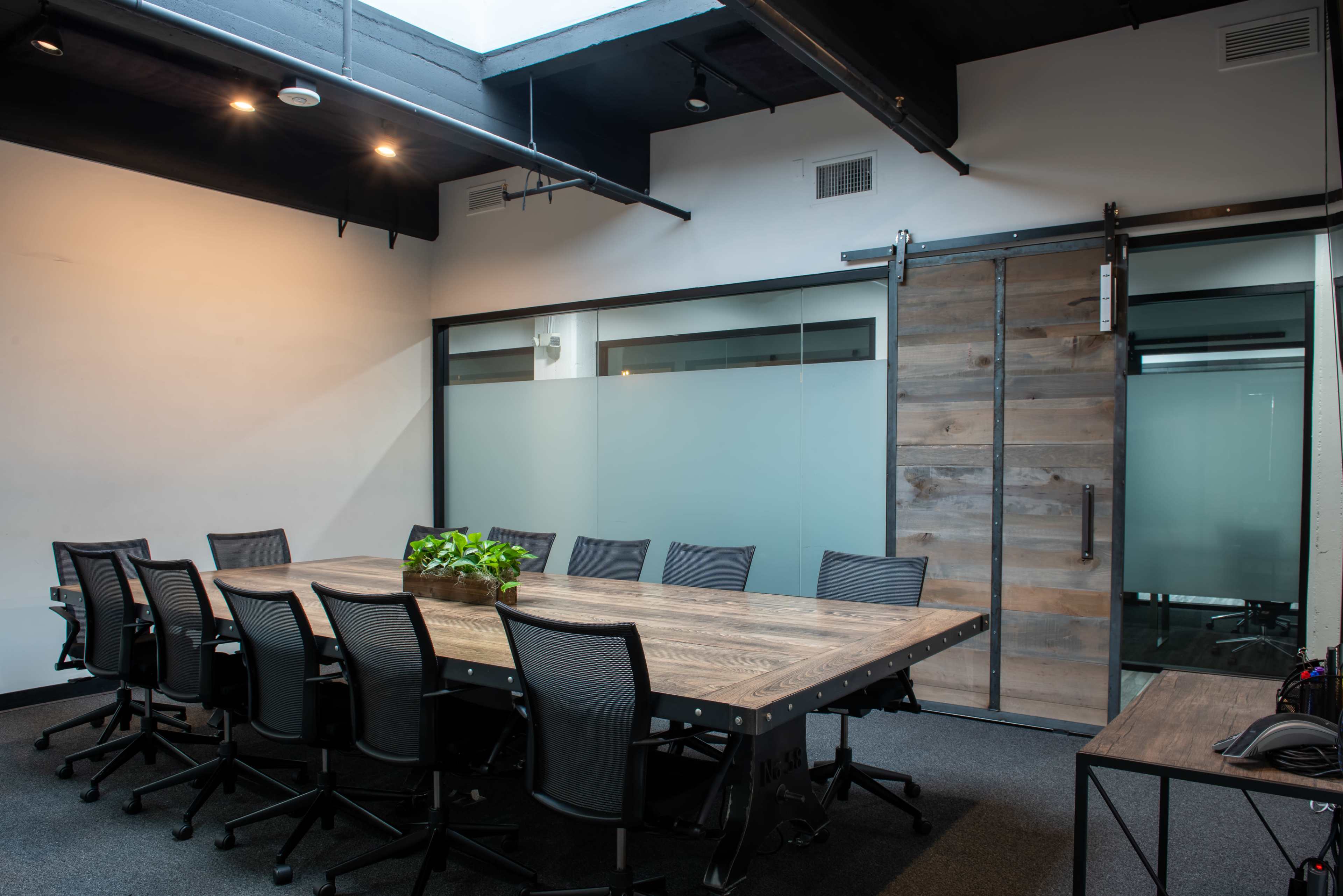 A conference room features a long wooden table surrounded by eight black chairs, with a sliding barn door and a plant centerpiece.