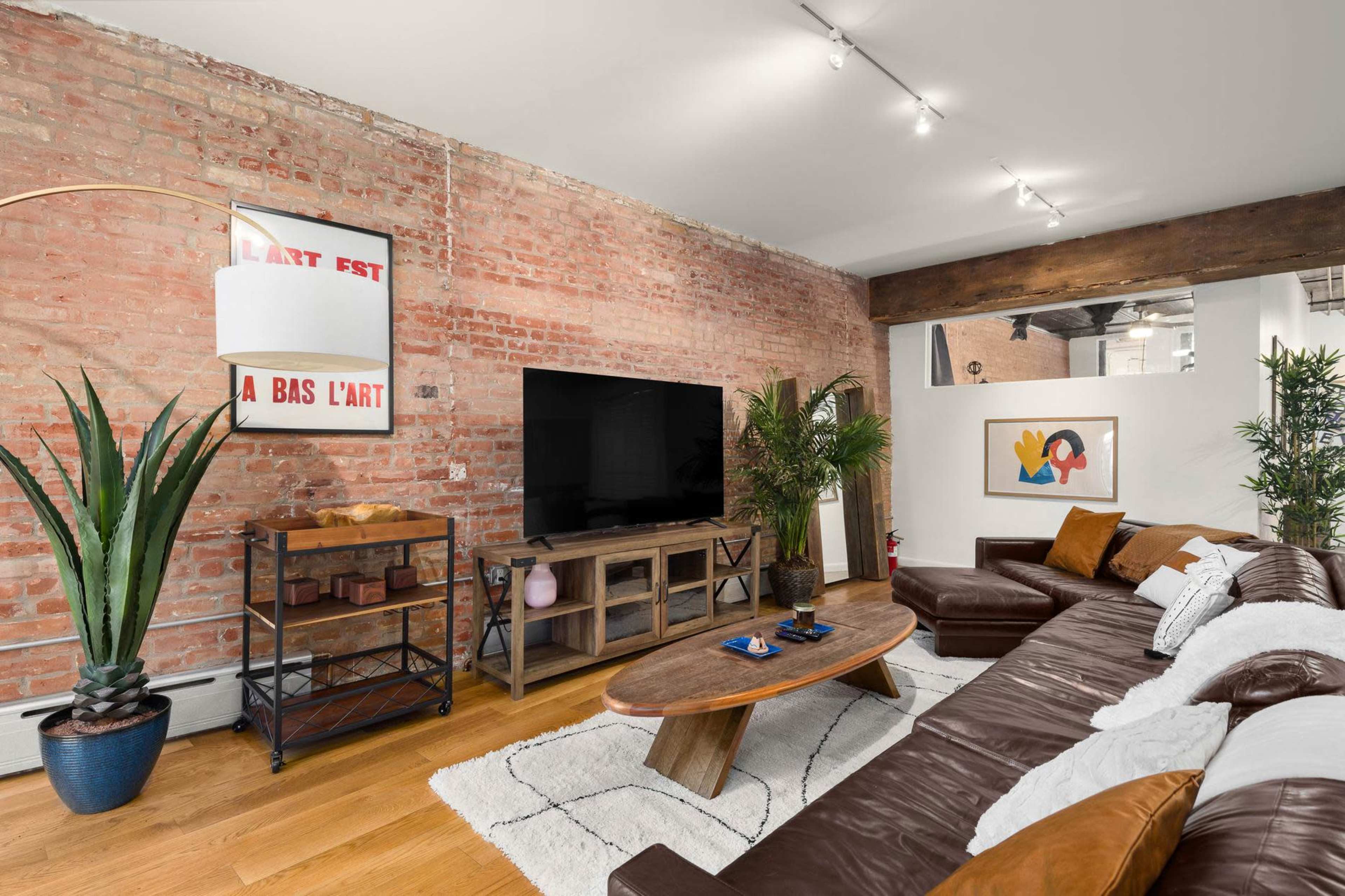 A cozy living room with exposed brick walls, a large sectional leather sofa, a wooden coffee table, and a TV mounted on the wall, surrounded by decorative plants and artwork.