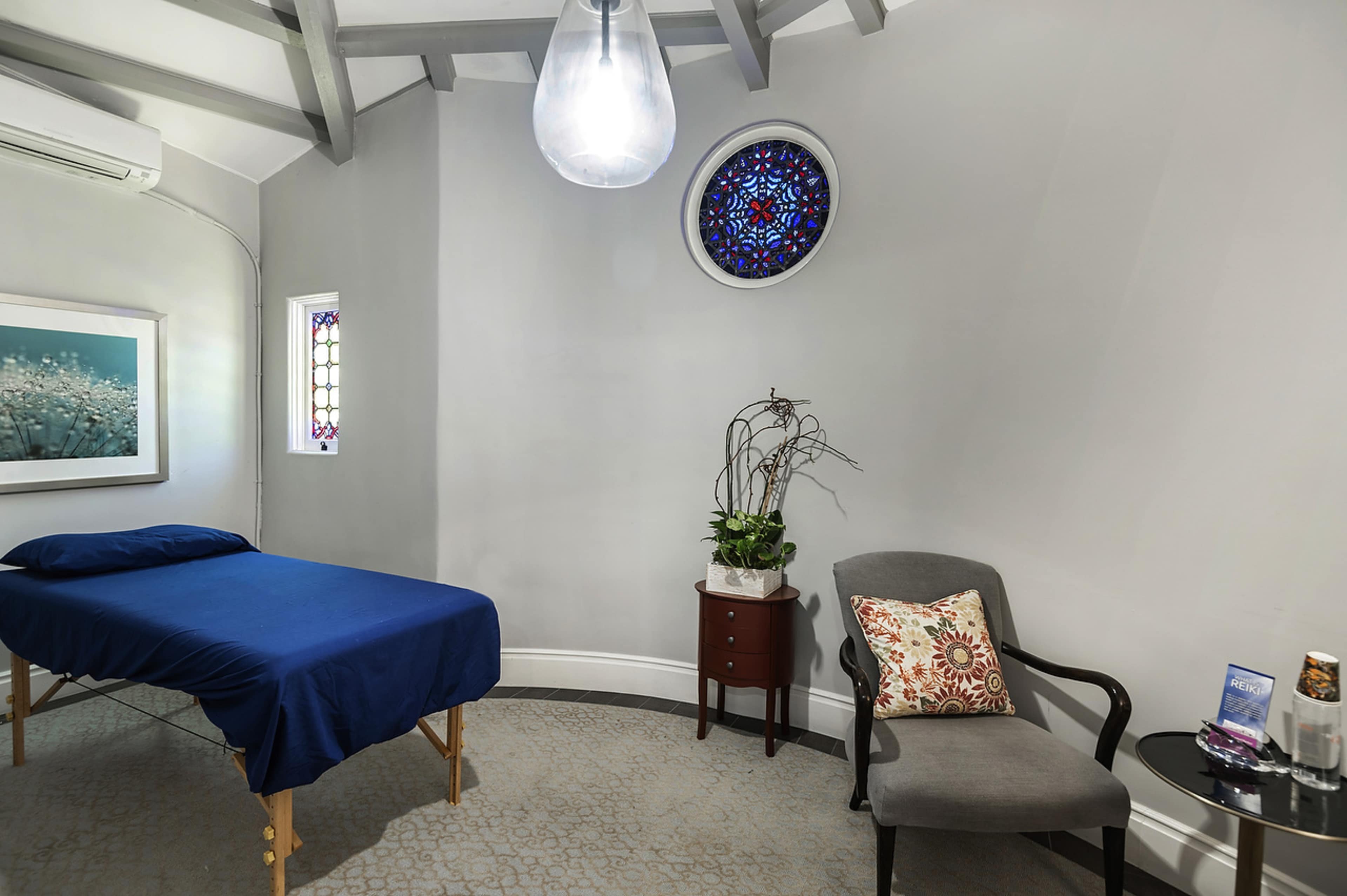 A small room features a massage table with a blue cover, a chair with a decorative pillow, and a small table with items on it, illuminated by a pendant light and a stained glass window.