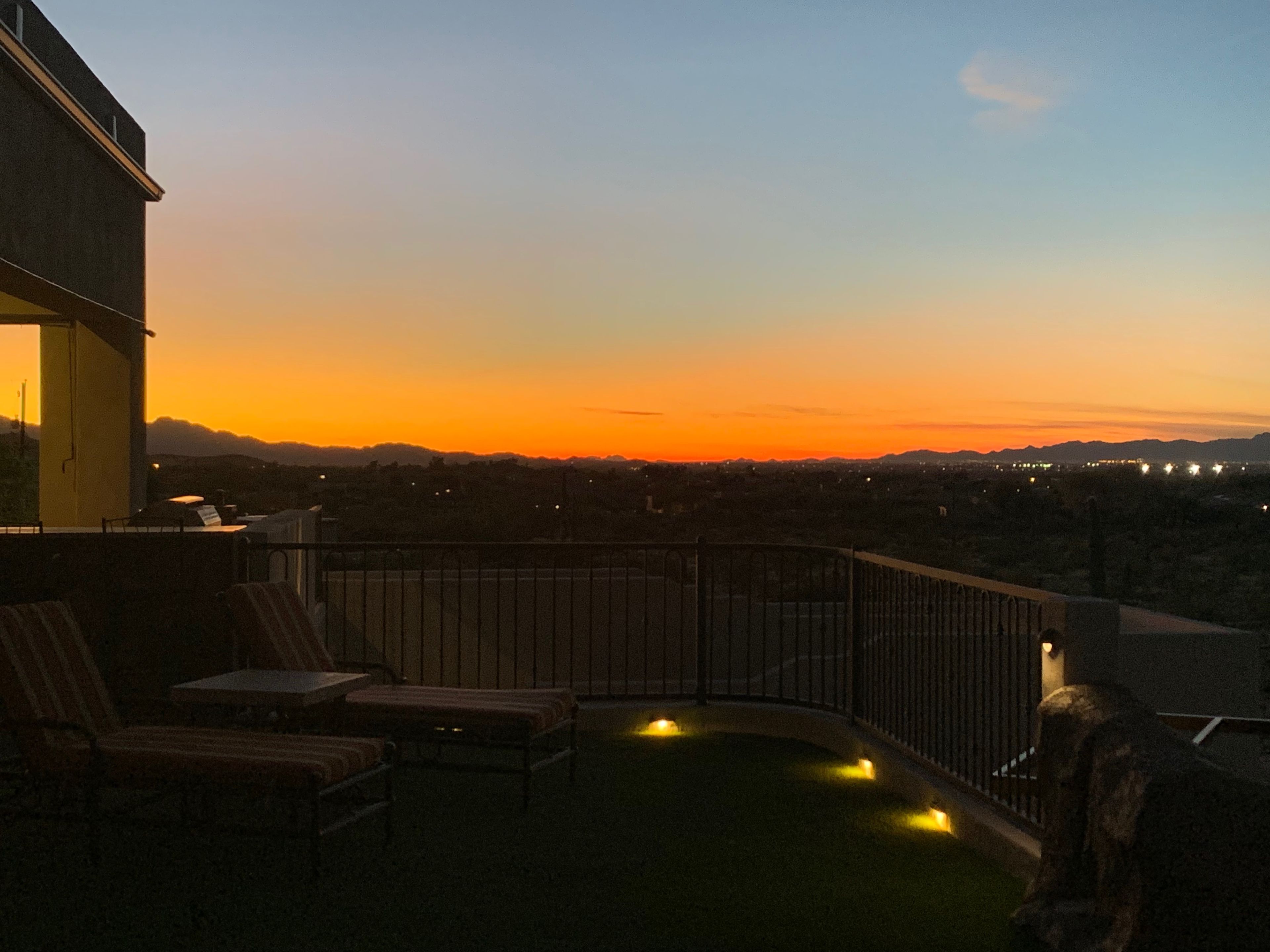 The image shows a rooftop terrace with striped lounge chairs overlooking a sunset that lights up the horizon and distant mountains.