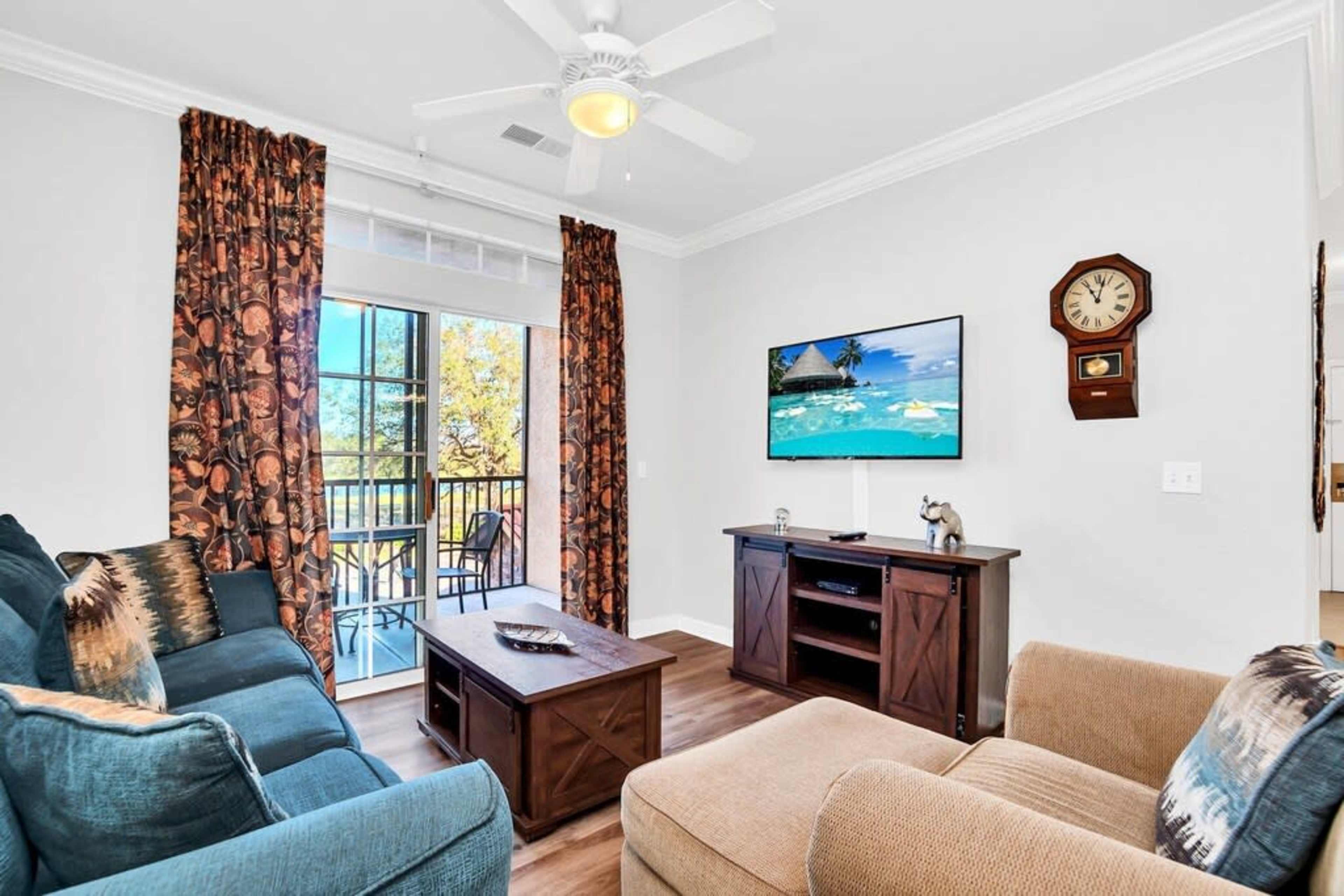 A living room with a blue sofa, a light brown coffee table, a television mounted on the wall, and a view of a balcony through large windows.