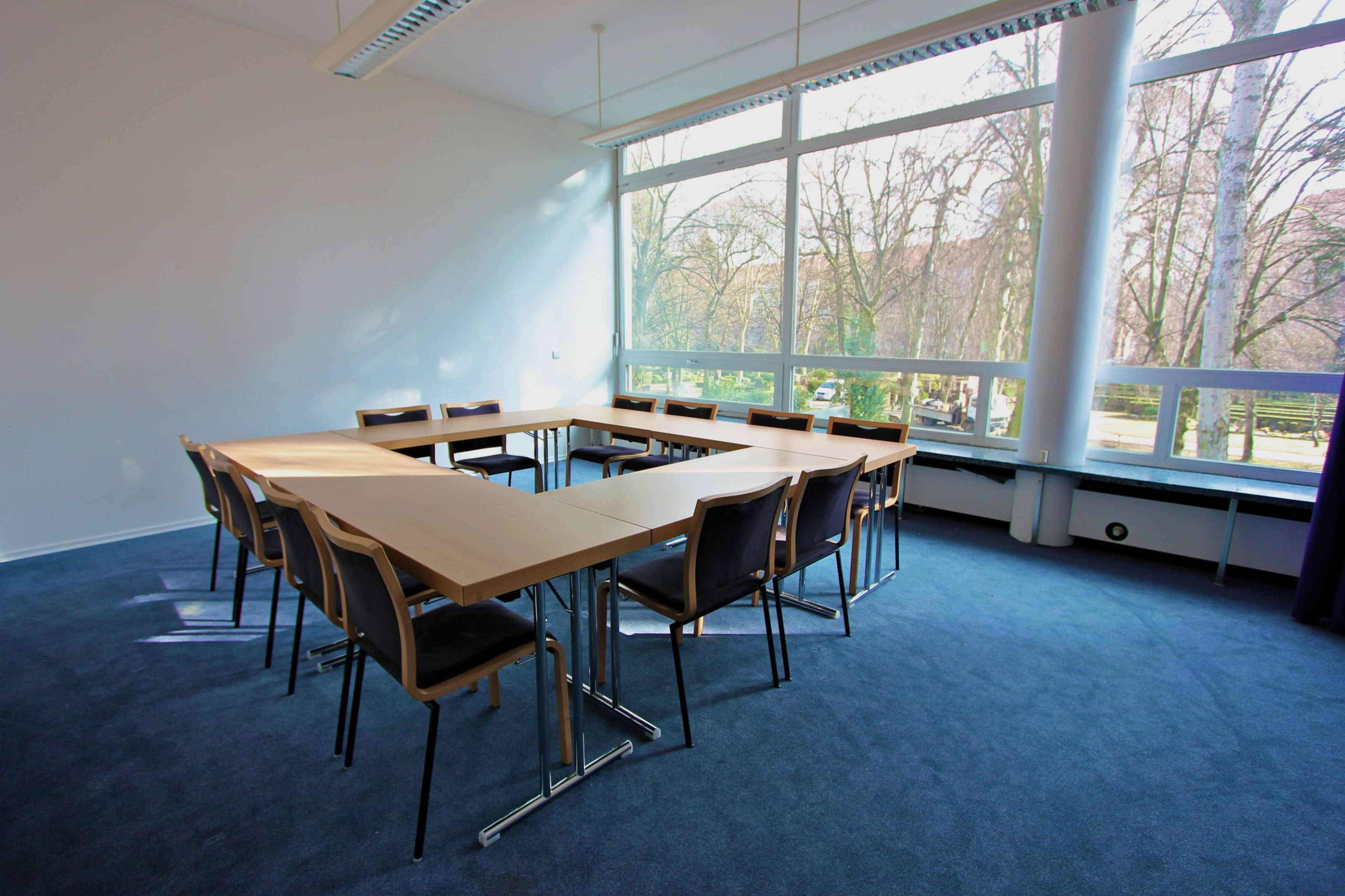 A meeting room features a large rectangular table surrounded by several chairs, with large windows providing natural light and a view of trees outside.