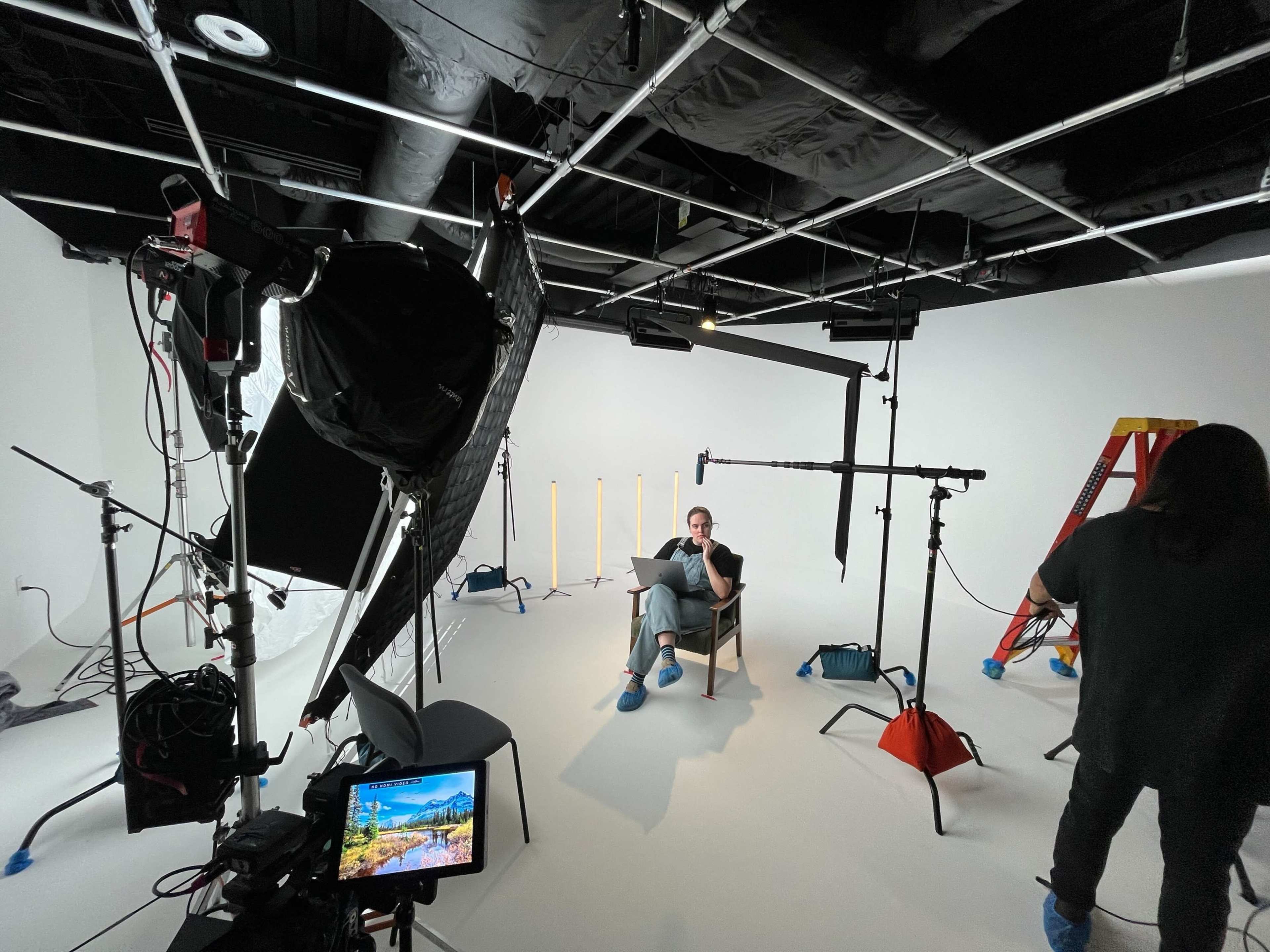 A person sits on a chair in a well-lit studio surrounded by lighting equipment and a ladder, while another individual moves in the background.