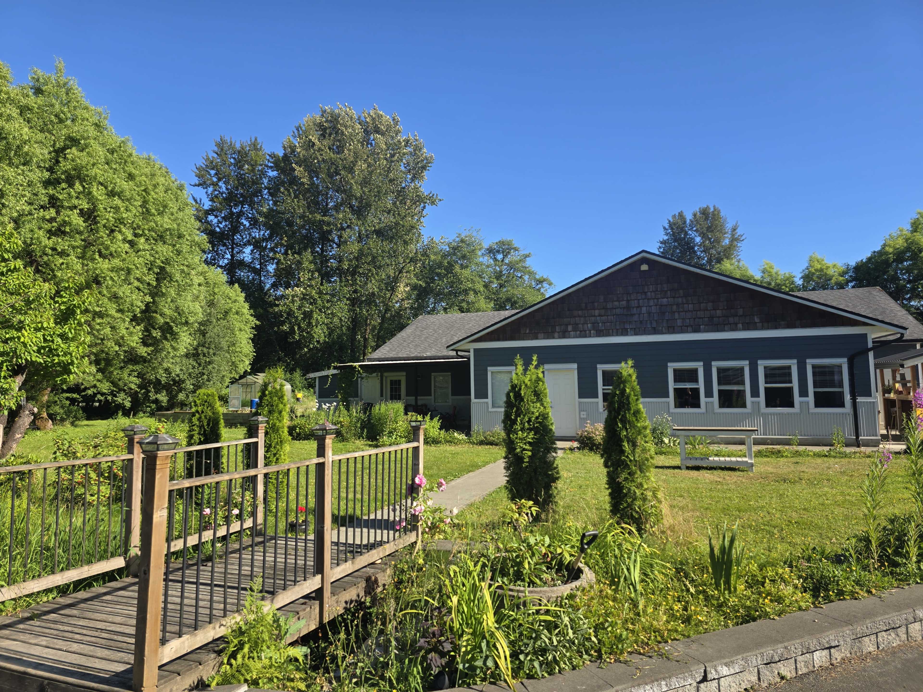 A single-story house with a dark roof and white trim is set amidst a lush green landscape, featuring a wooden footbridge and well-maintained garden areas.