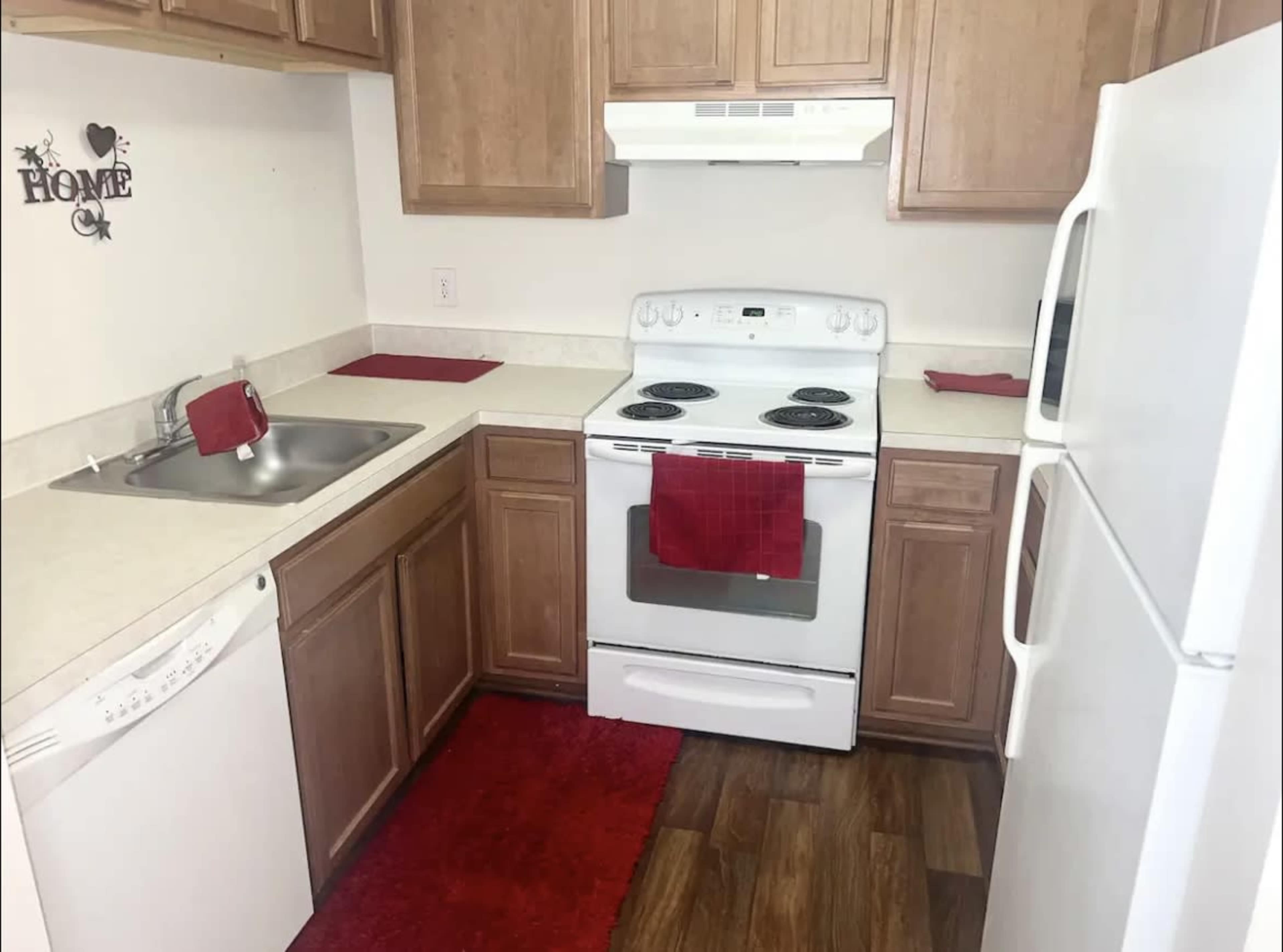 A kitchen with wooden cabinets, a white stove, a dishwasher, and a red dish towel on the counter, along with a matching red rug on the floor.