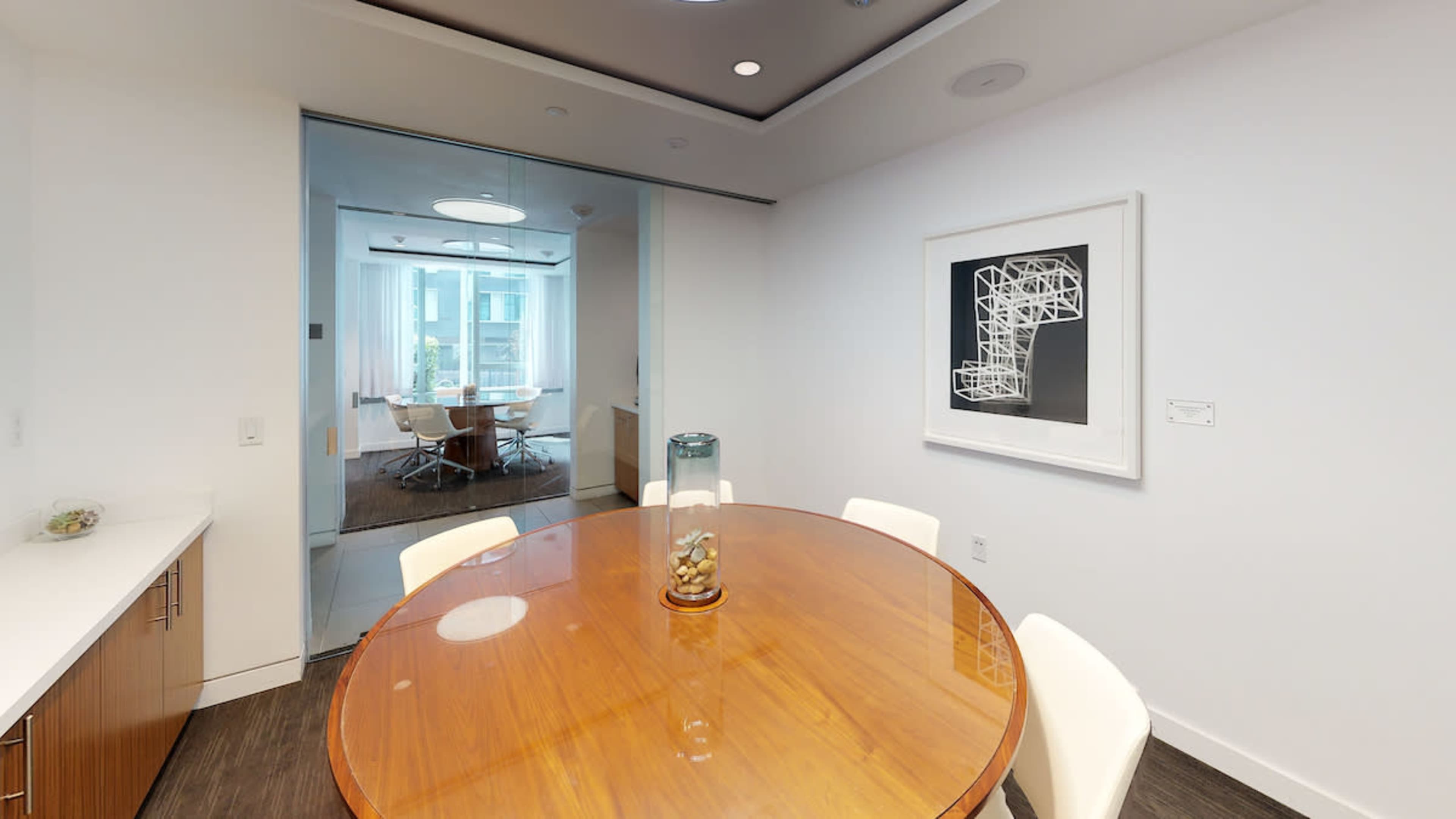 A round wooden dining table surrounded by white chairs is positioned in a brightly lit room with modern decor and glass walls.