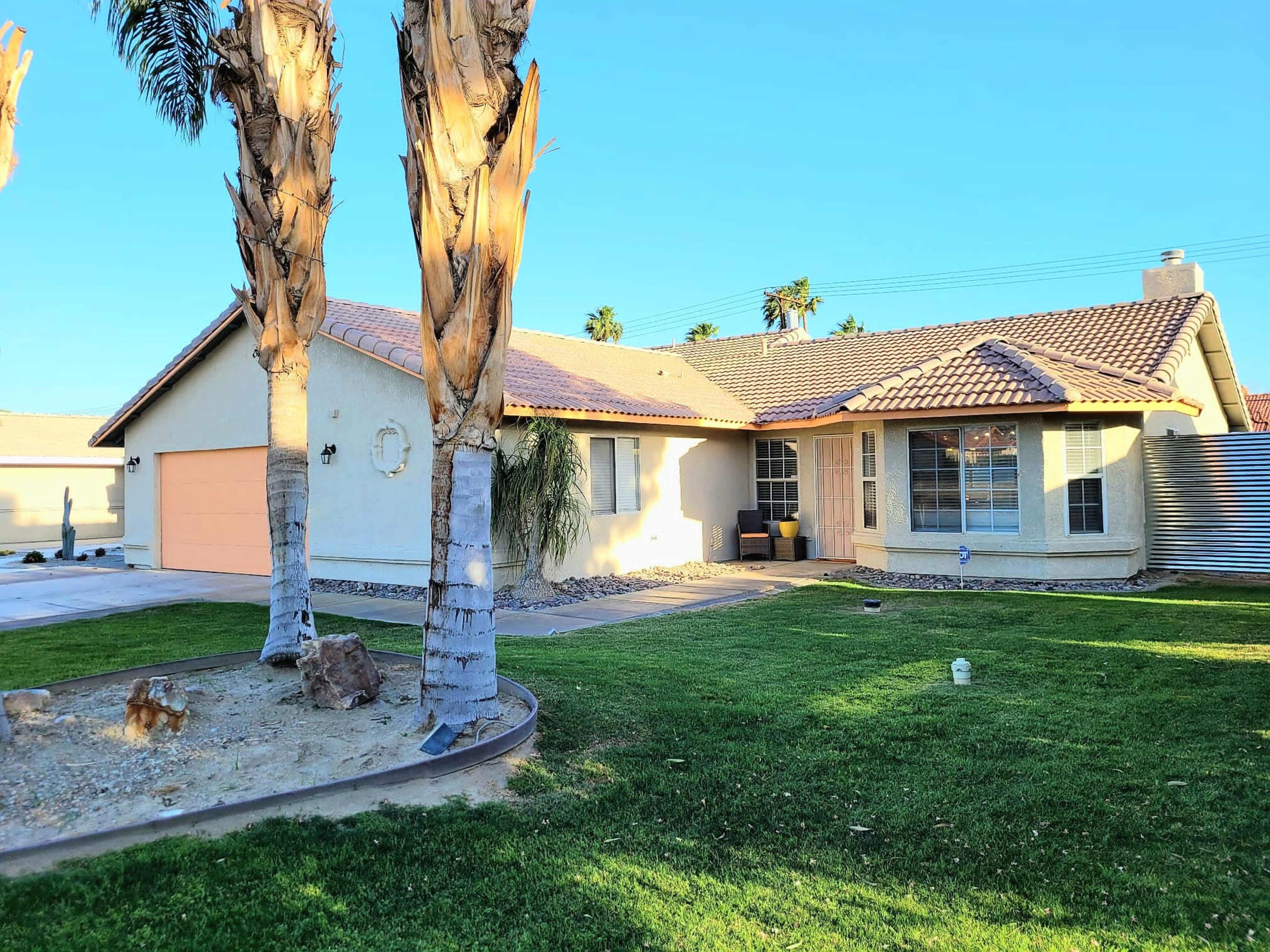 A single-story house with a beige exterior and a tiled roof sits in a yard flanked by palm trees.
