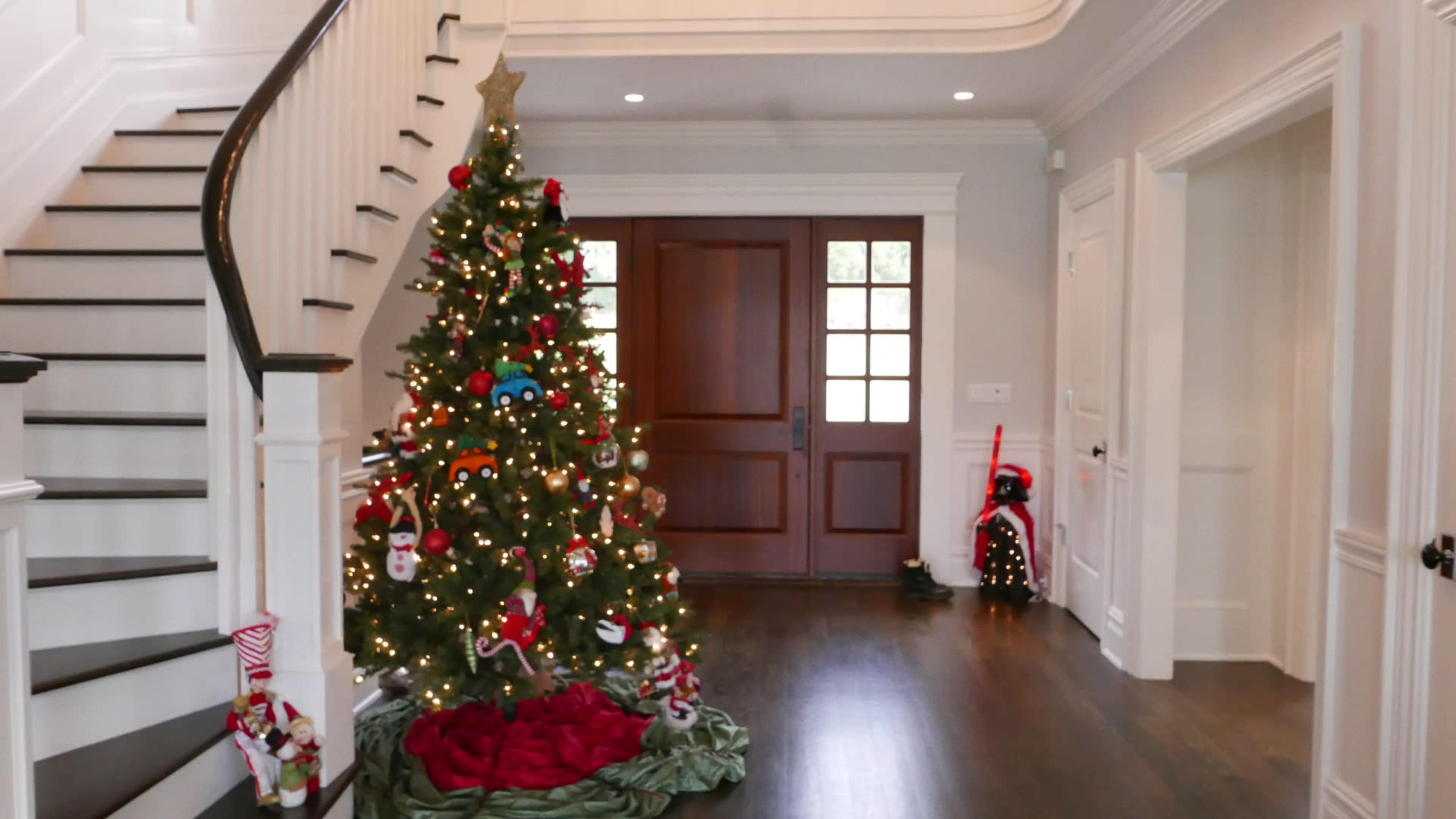 A decorated Christmas tree stands in a well-lit foyer next to a double wooden door, with a staircase visible in the background.