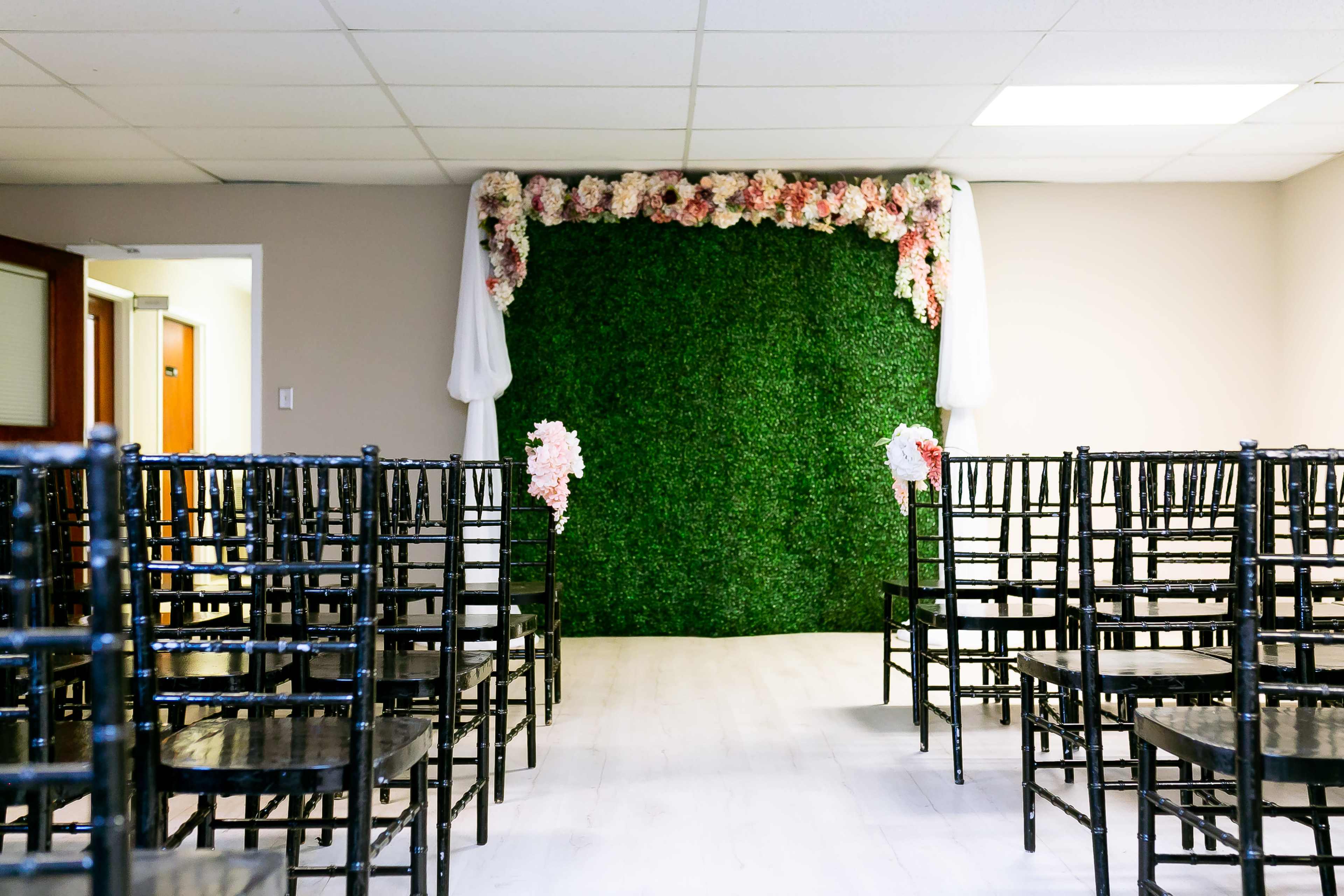 The image shows a wedding ceremony setup with rows of black chairs facing a floral and greenery backdrop, adorned with white drapery.