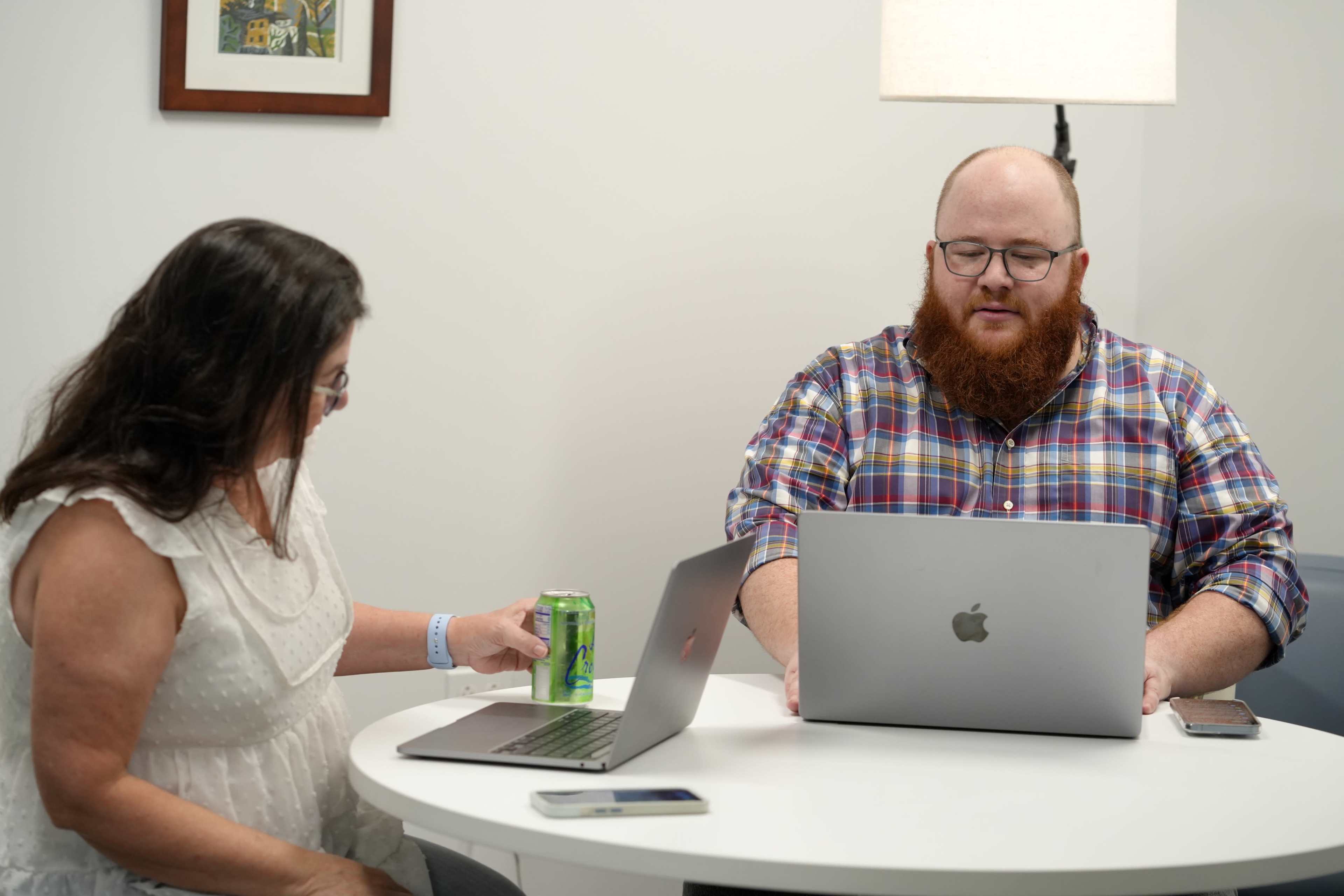 A woman sits at a table with a can of soda while a man in a plaid shirt works on a MacBook.