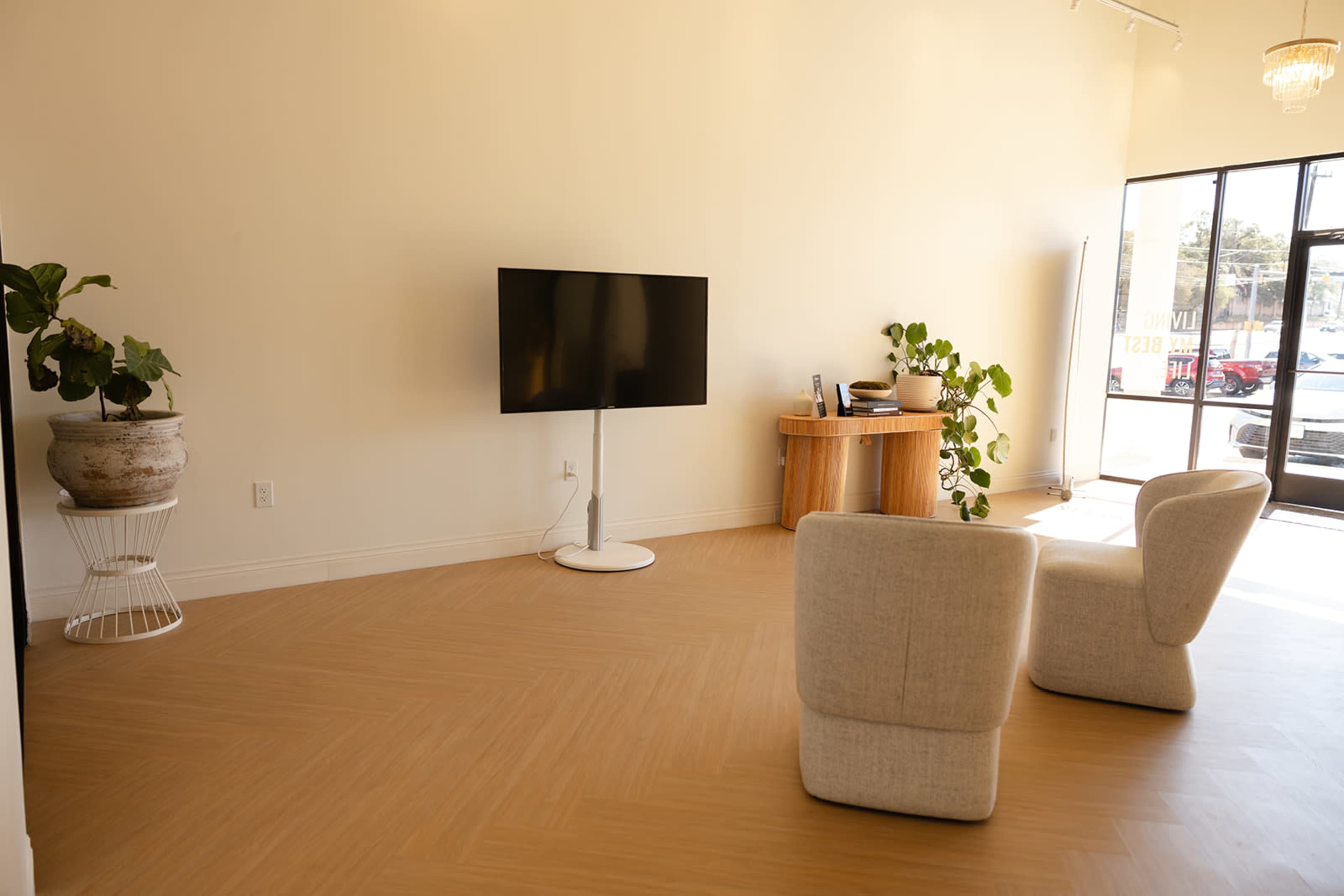 The image shows a minimally furnished interior space with a television on a stand, a round table, and two armchairs, illuminated by natural light from a large window.