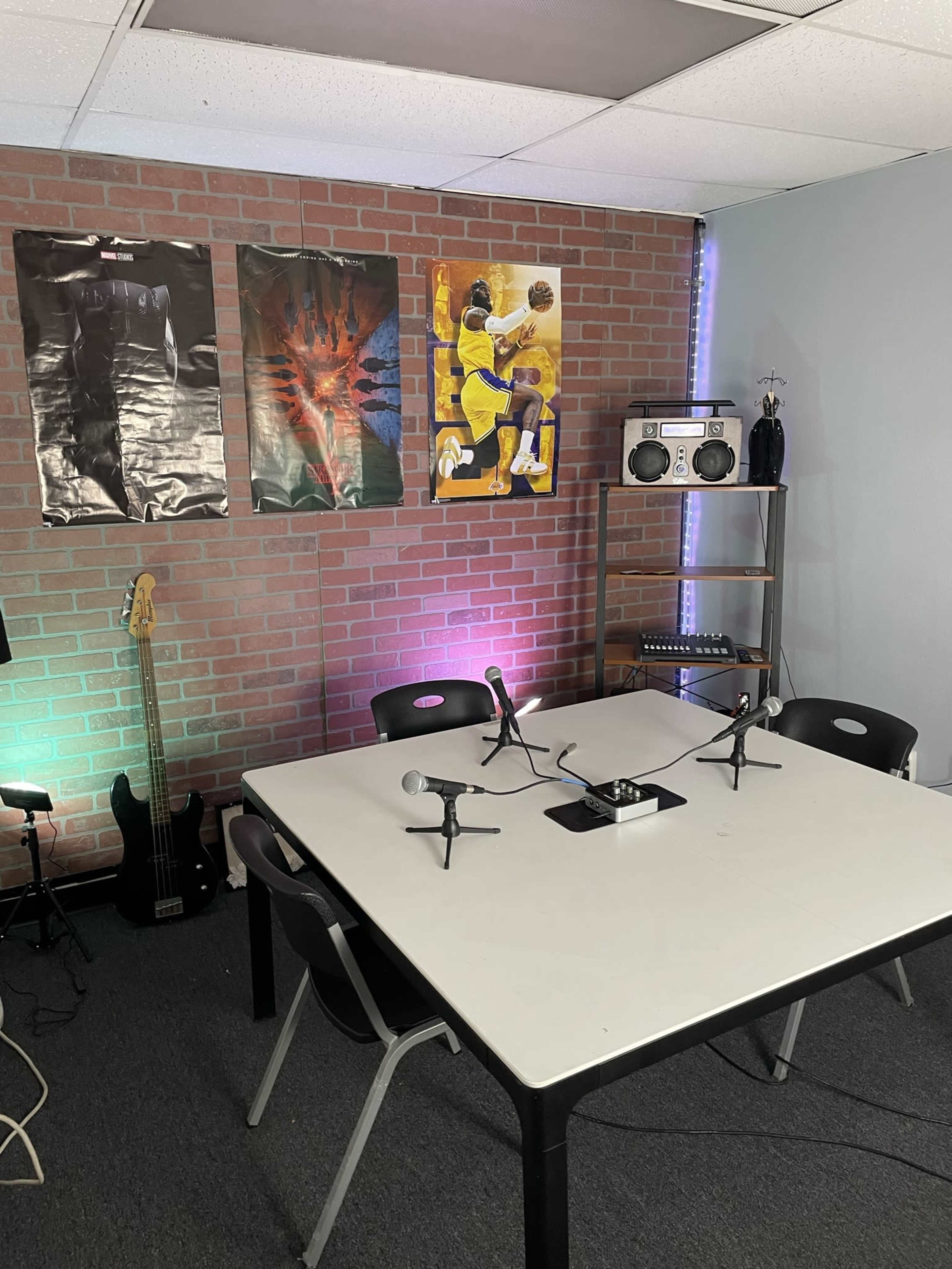 A small recording or meeting space with a table at the center, equipped with microphones, a boombox, and three posters on a brick wall, alongside a guitar and a shelf.